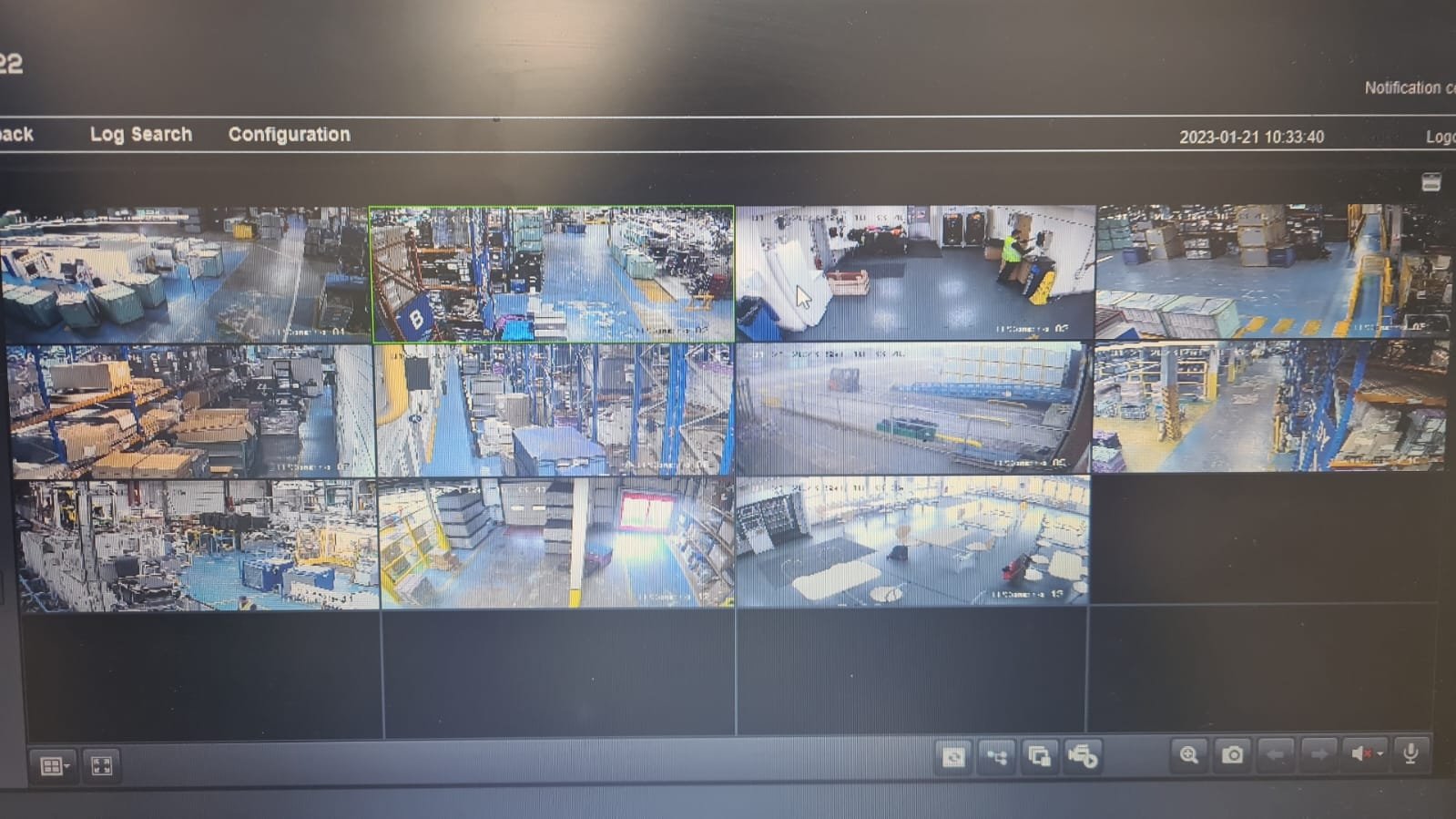 Security camera monitor displaying multiple camera feeds of a warehouse, showing aisles with shelves, boxes, forklifts, and workers.