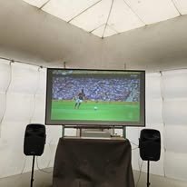 Flat-screen TV showing a soccer match, on a table with two speakers, inside a white tent