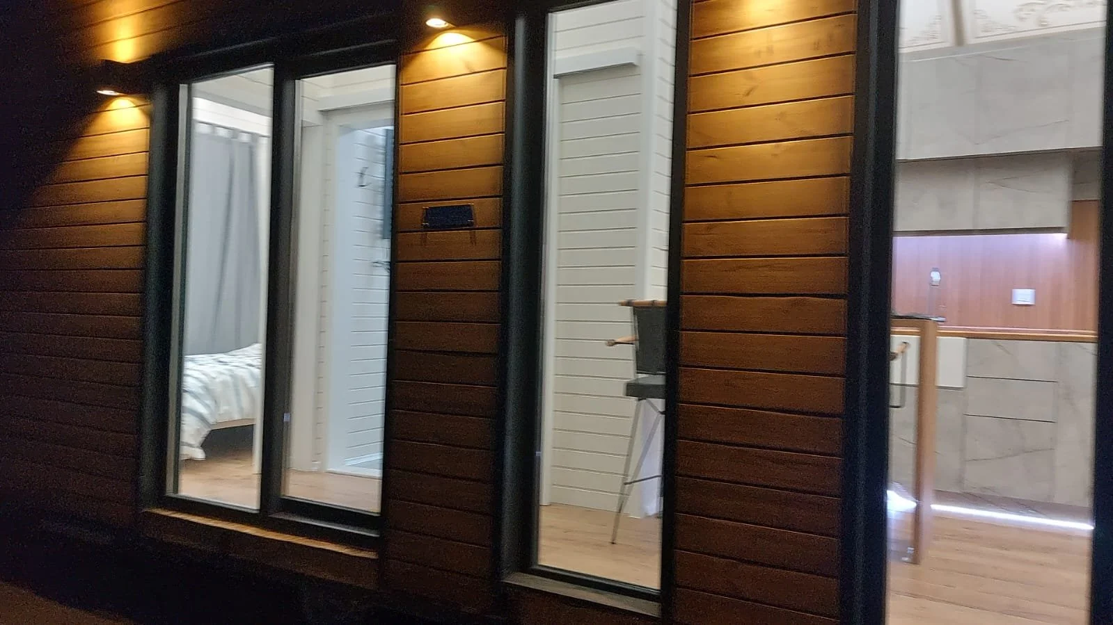 View of an outdoor balcony with sliding glass doors showing a bedroom on the left and an interior kitchen on the right, with warm lighting and paneled wood exterior walls.