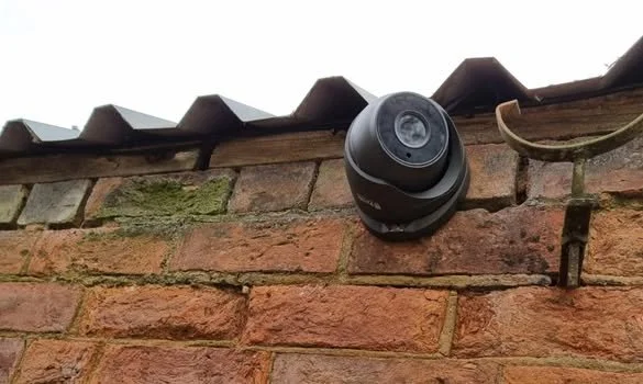 Security camera mounted on a brick wall beneath a wooden roof edge with metal roofing shingles.