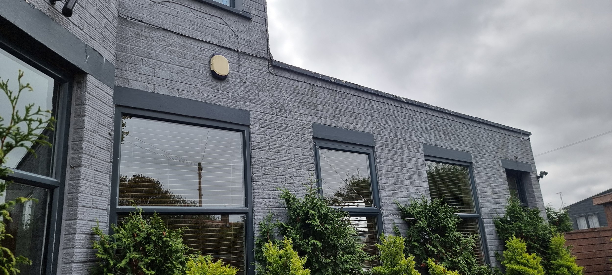 Gray brick building with three large windows and greenery in front, cloudy sky overhead.