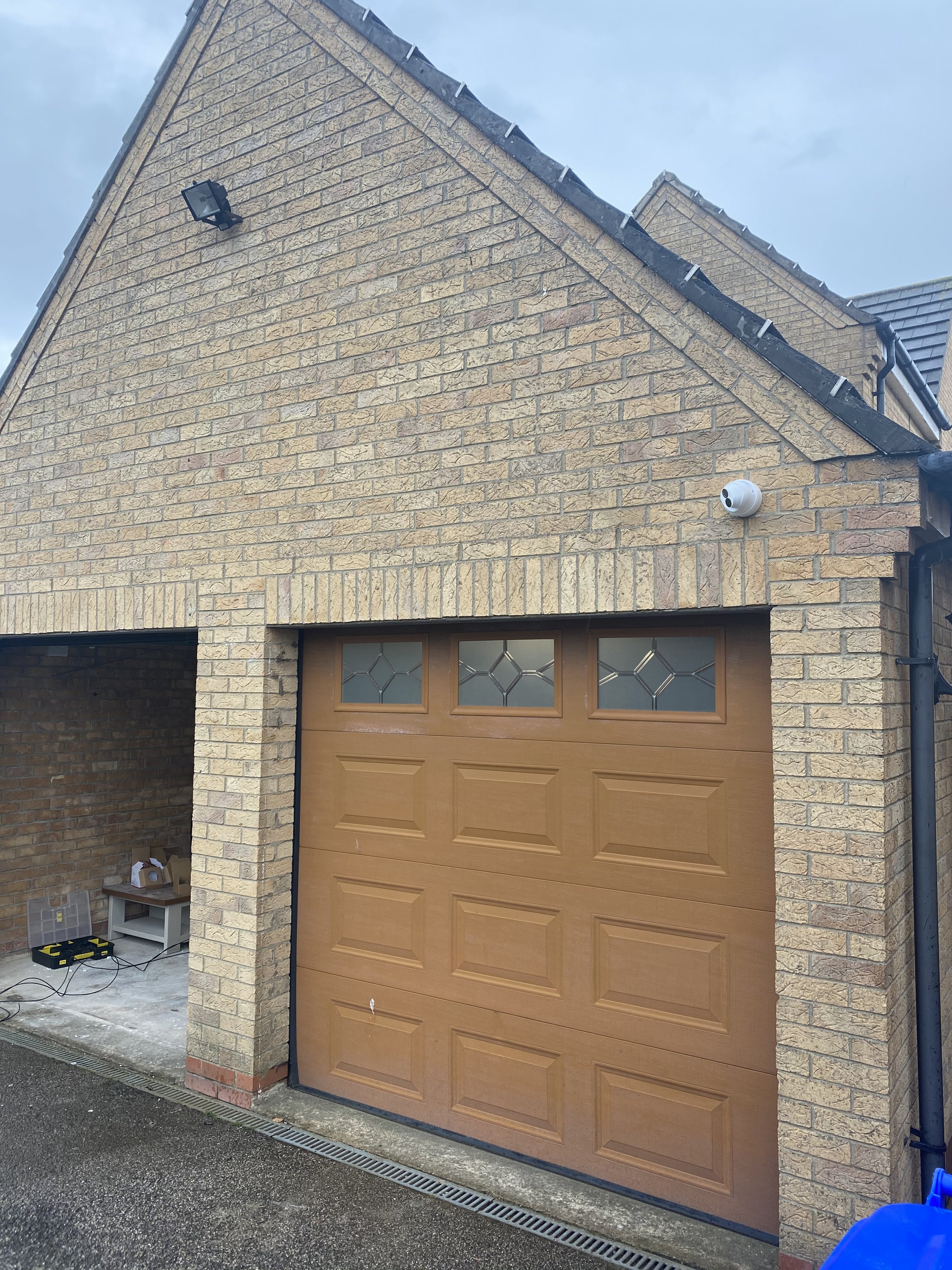 A brick house with a wooden garage door, security camera, floodlight, and construction tools on the ground.