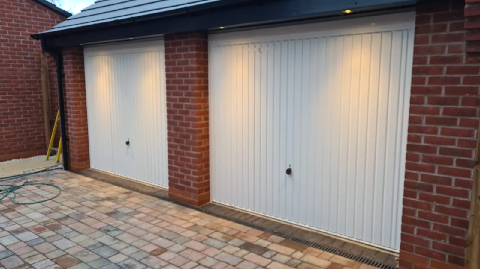 Two white tilting garage doors installed on a brick garage with some construction tools and wires nearby.