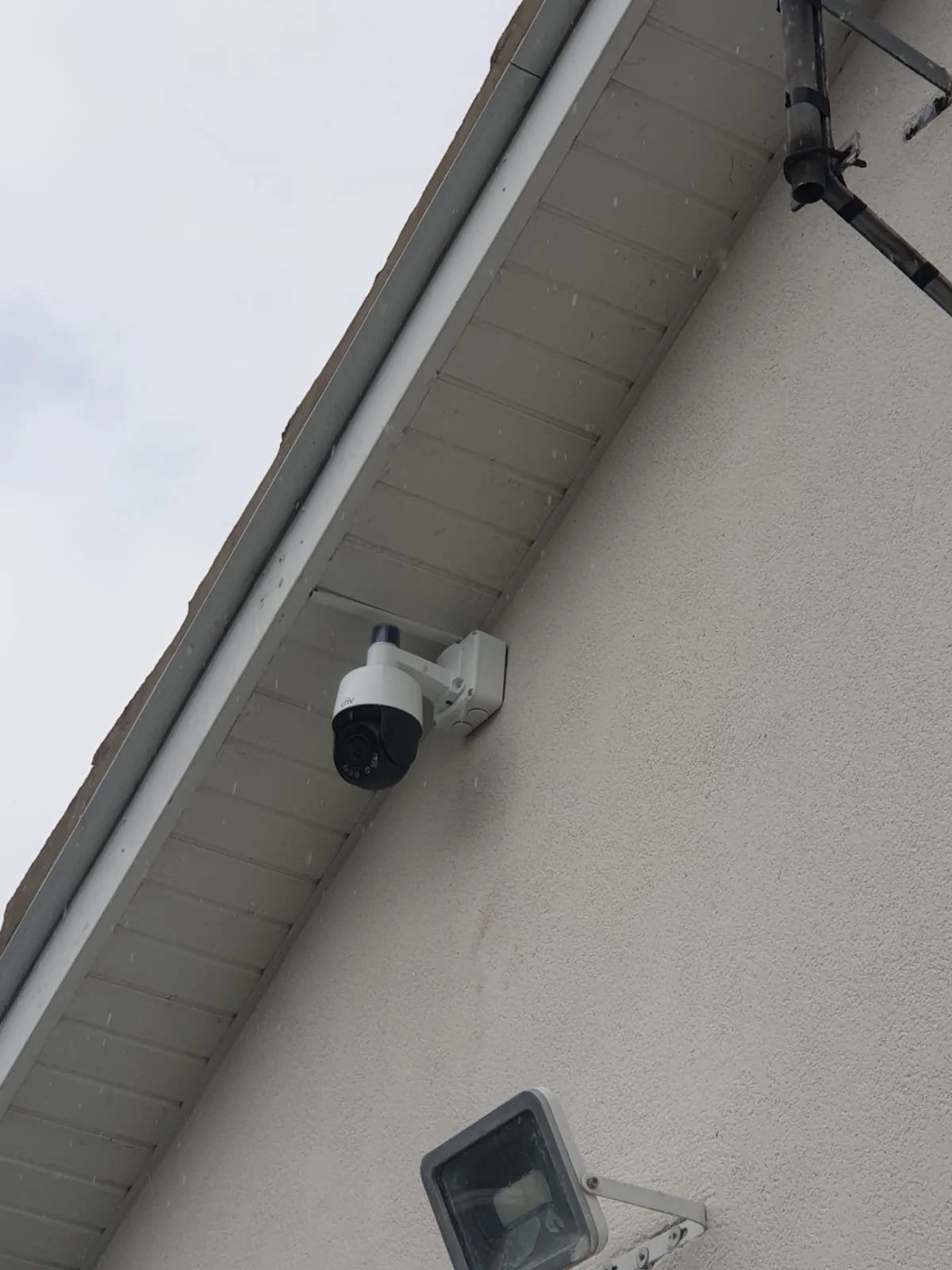 Security camera mounted under eaves on a house exterior, with an outdoor light fixture nearby.