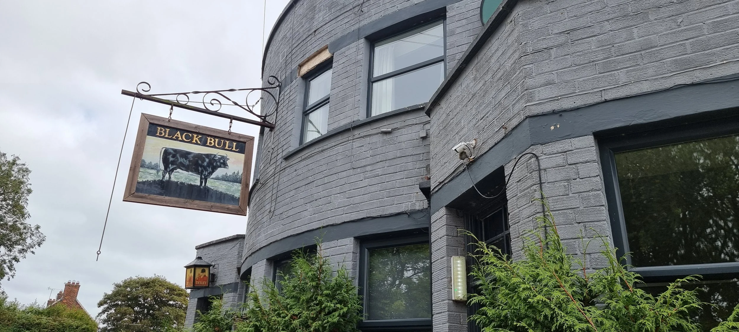 Gray brick building with a sign that reads 'Black Bull' showing a black and white bull. The building has three visible windows, some greenery in front, and the sky is cloudy.