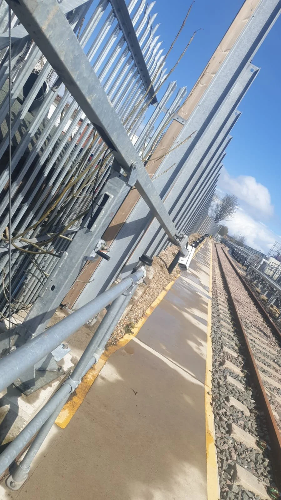 View of a railway track with a concrete pedestrian pathway beside it, with a metal fence and electric cables along the pathway, under a partly cloudy sky.