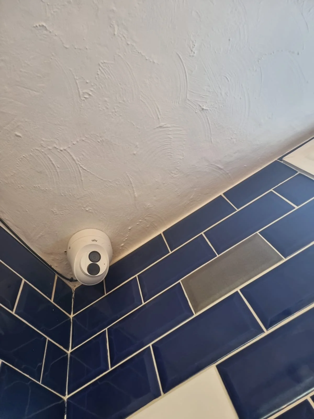 A white security camera mounted on the corner of a wall with blue tile and white grout, and a textured white ceiling.