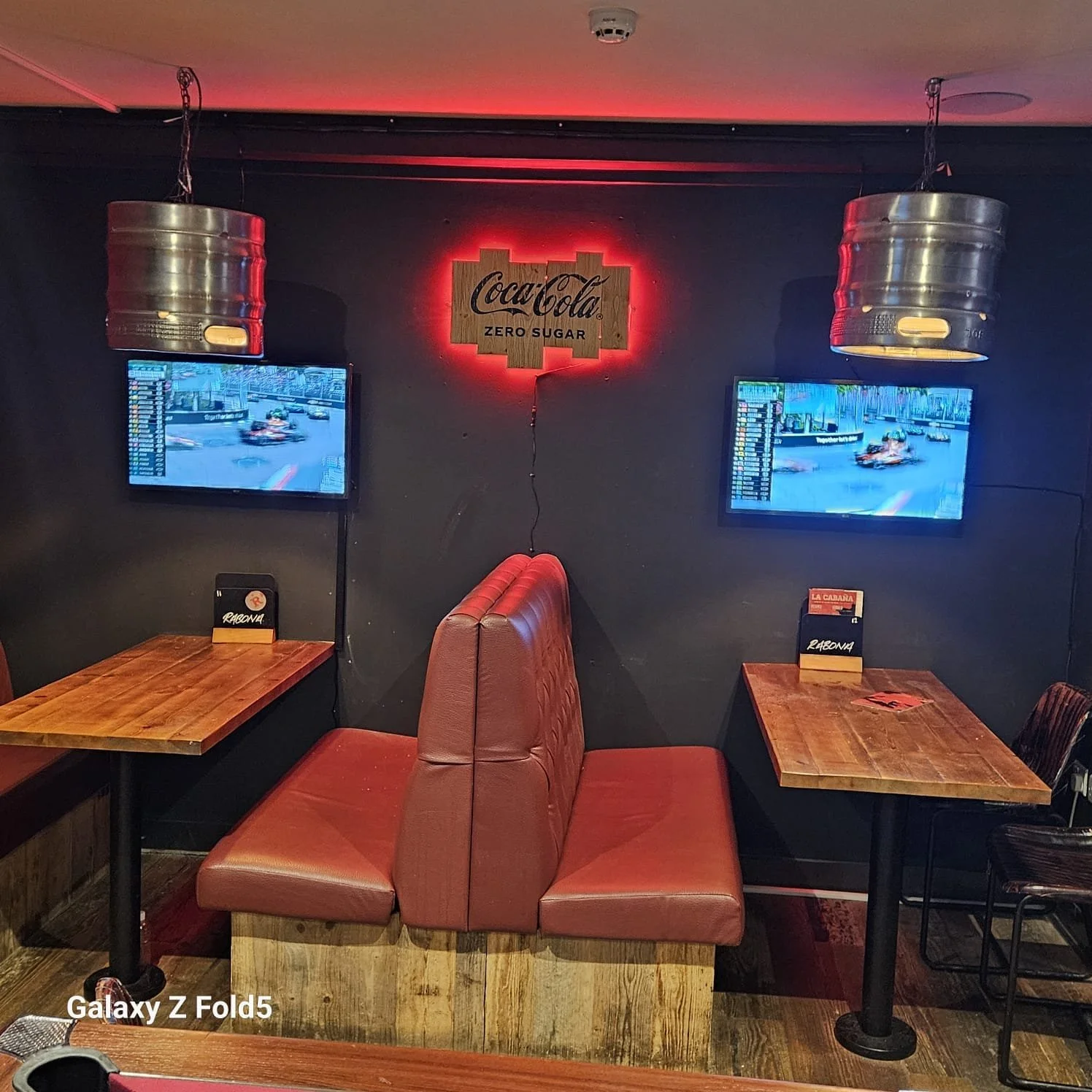 A cozy restaurant or bar seating area with wooden tables, red upholstered booths, two wall-mounted televisions showing a racing event, a Coca-Cola Zero Sugar sign with red backlight, and metallic pendant lights hanging from the ceiling.