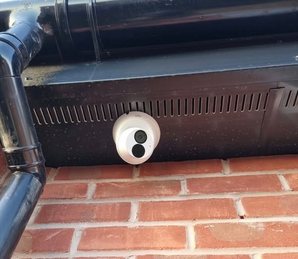 Security camera mounted below a black vent on a brick wall.