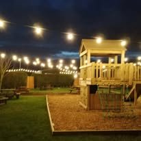 Outdoor area at night with string lights, a wooden deck with a railing, a small swing set, and a grassy yard.
