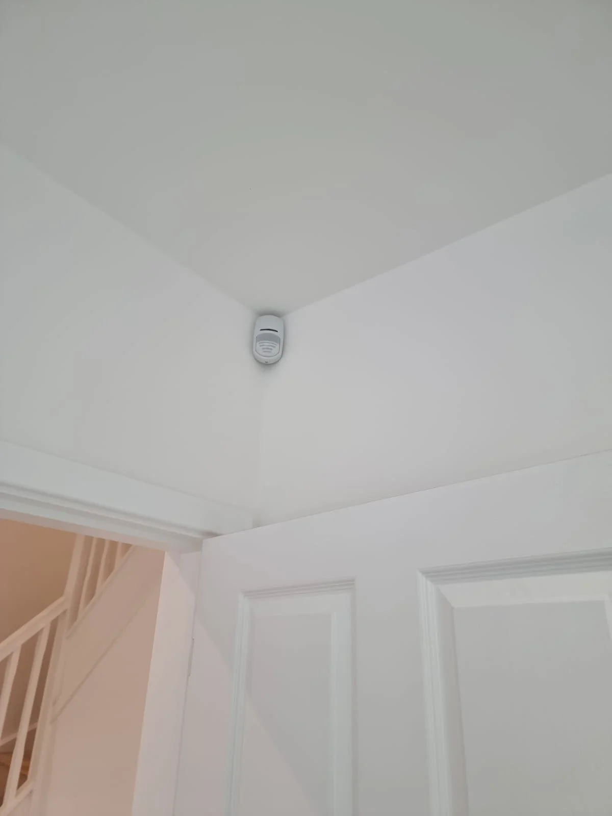 Smoke detector mounted on white ceiling in a corner of a room near a white door.