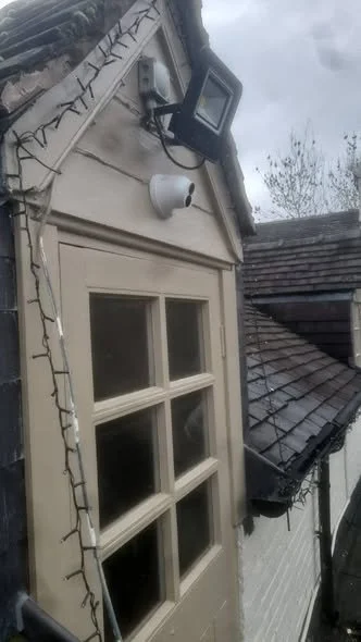 Exterior of a house with a window, a security camera, and Christmas lights on the wall, with a cloudy sky in the background.