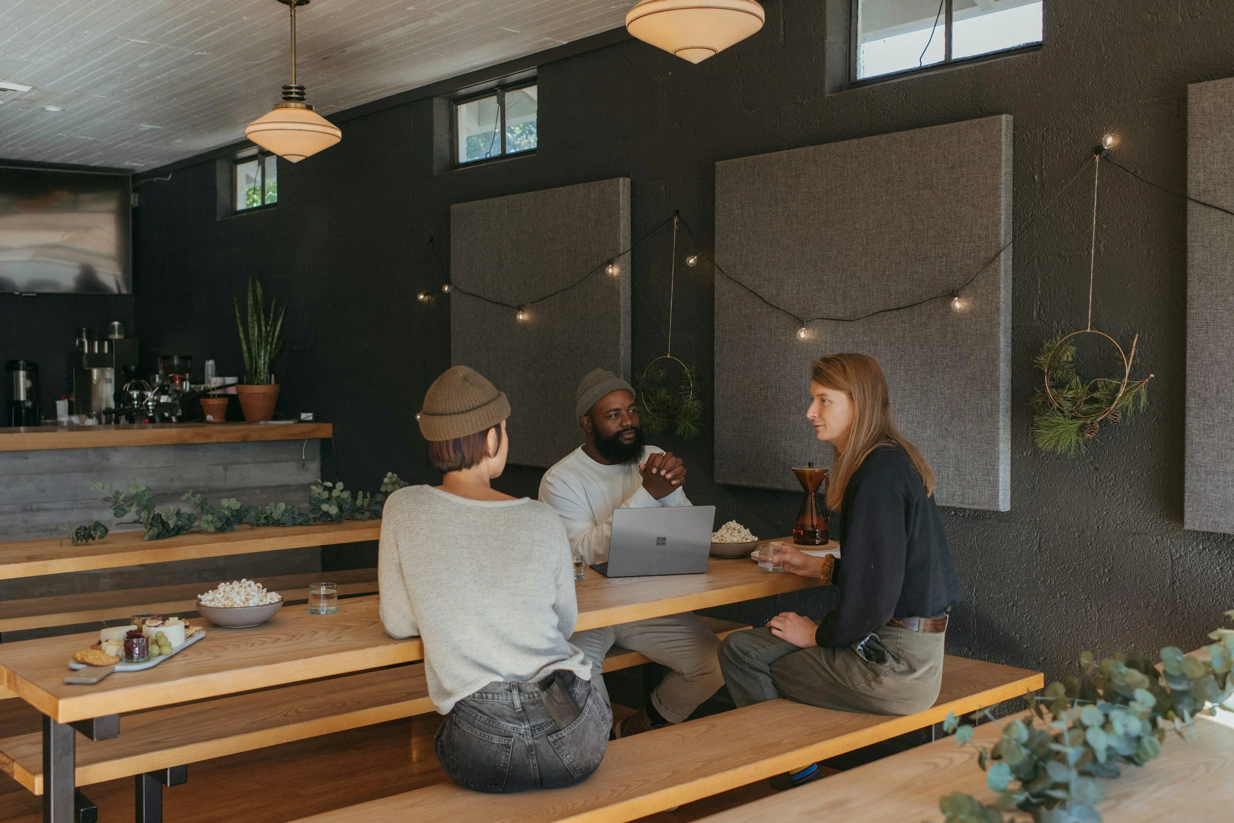 Three people are sitting and standing at a wooden table in a coffee shop, engaging in conversation. One person is using a laptop, and there are plates of desserts on the table. The background features a dark textured wall, hanging string lights, and a small window. The setting appears cozy and modern.