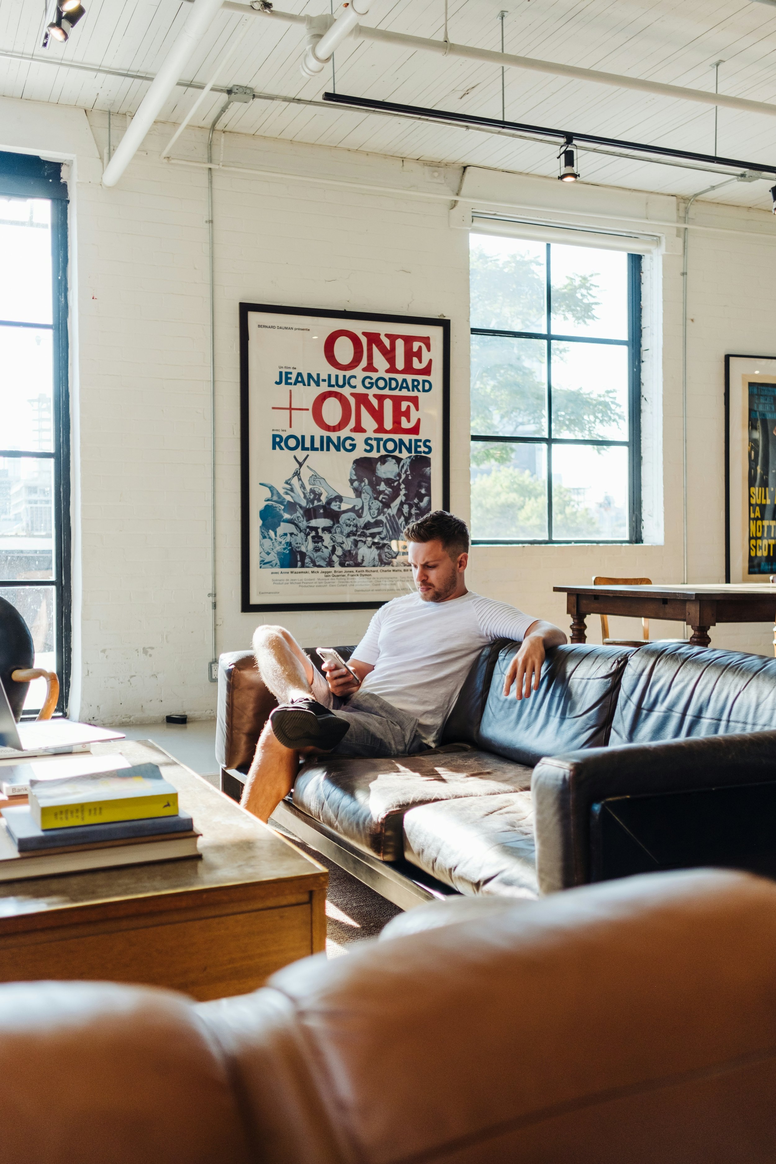 A man with short dark hair and a beard sitting on a brown leather sofa in a room with white brick walls and large windows, looking at his phone. There are posters on the wall, including one that says 'ONE + ONE' with images of The Rolling Stones.