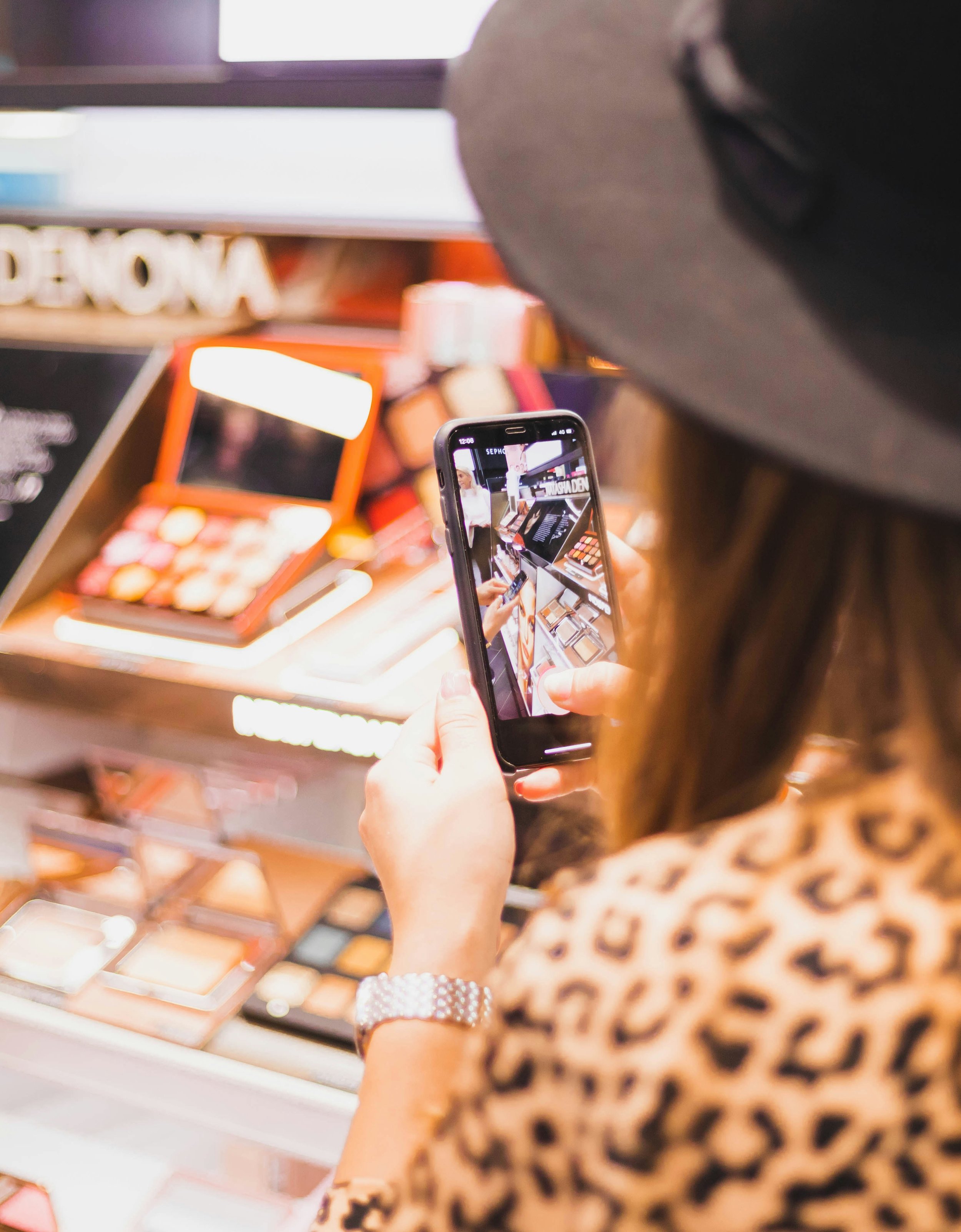 Person shopping for makeup products in a cosmetic store, taking a photo with smartphone, with makeup palettes on display in the background.