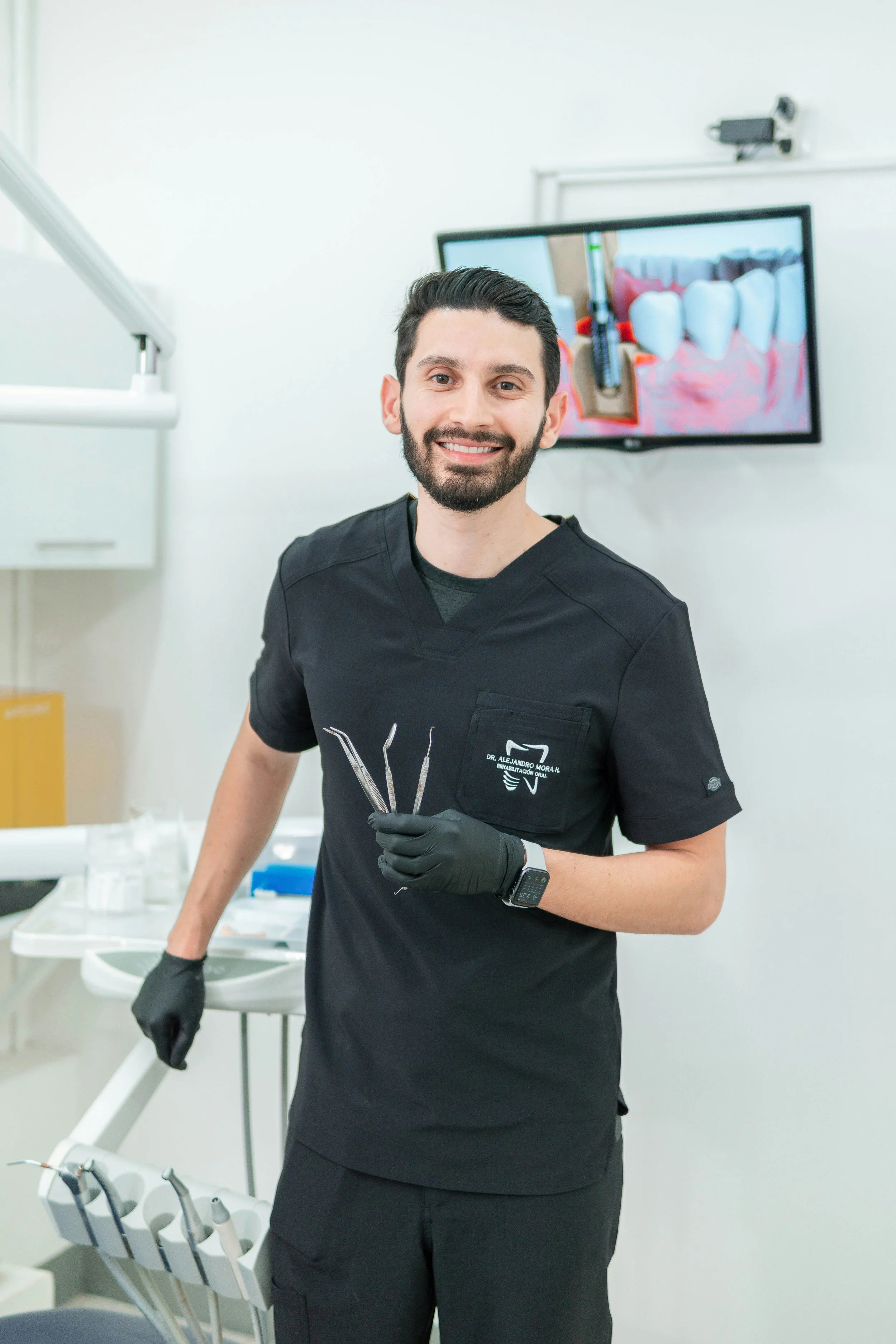 A smiling male dentist holding dental tools in a dental clinic, with a monitor displaying teeth and dental equipment in the background.