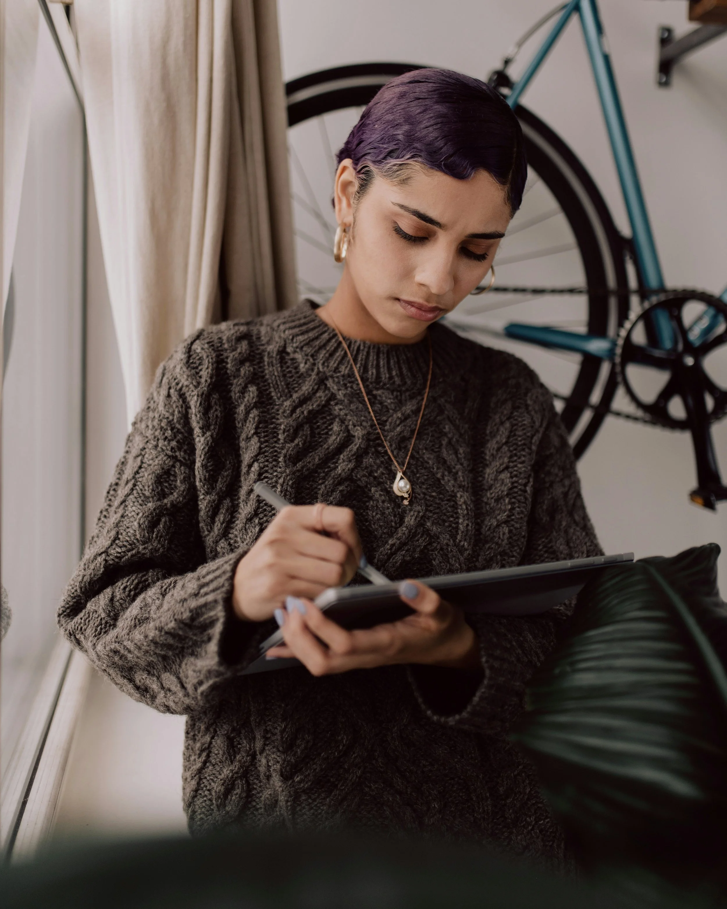 A young woman with short dark hair and earrings is looking down at a tablet, holding a stylus, sitting near a bicycle.