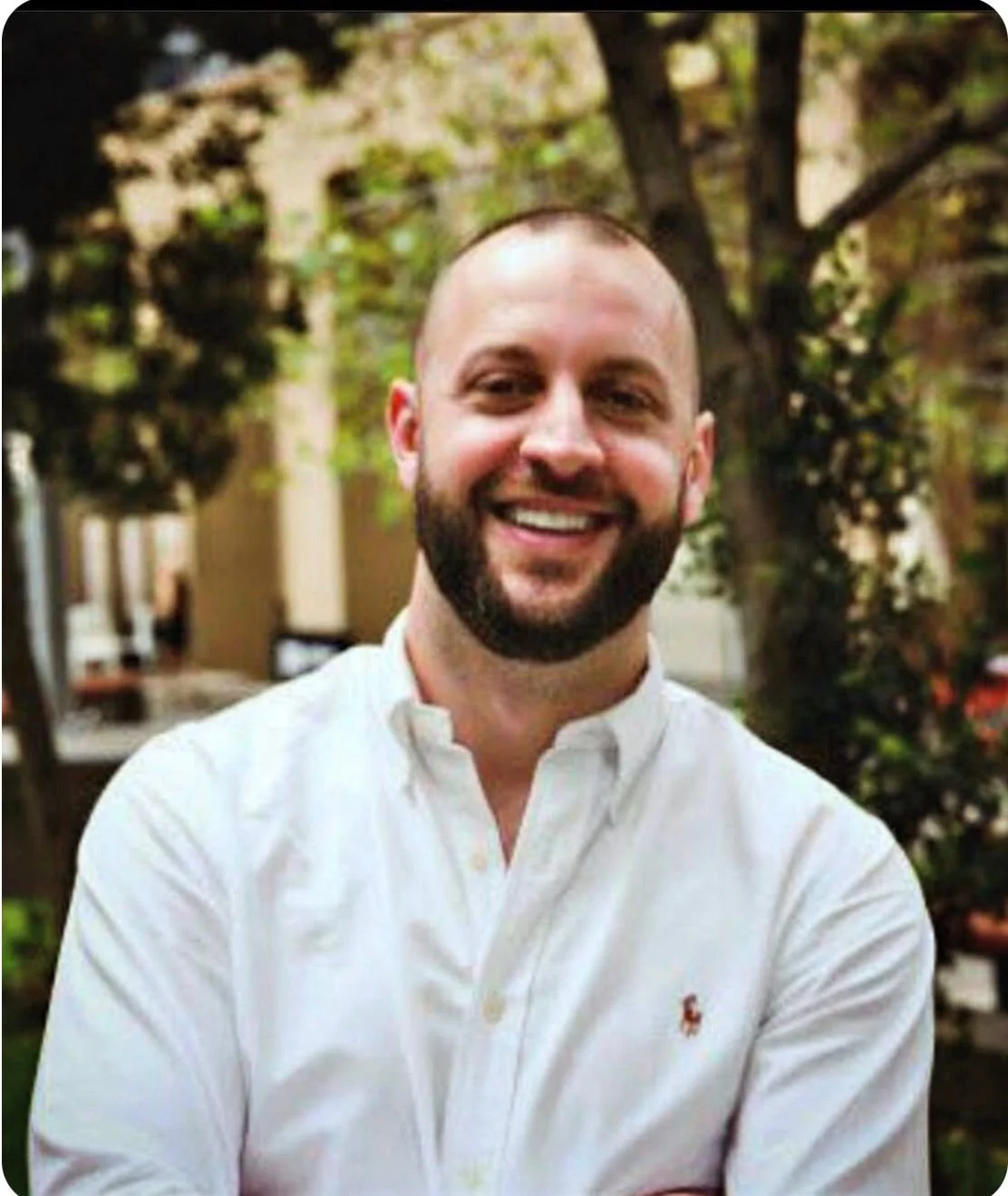 A smiling man with a beard wearing a white collared shirt outdoors in a garden or park with trees and plants in the background.
