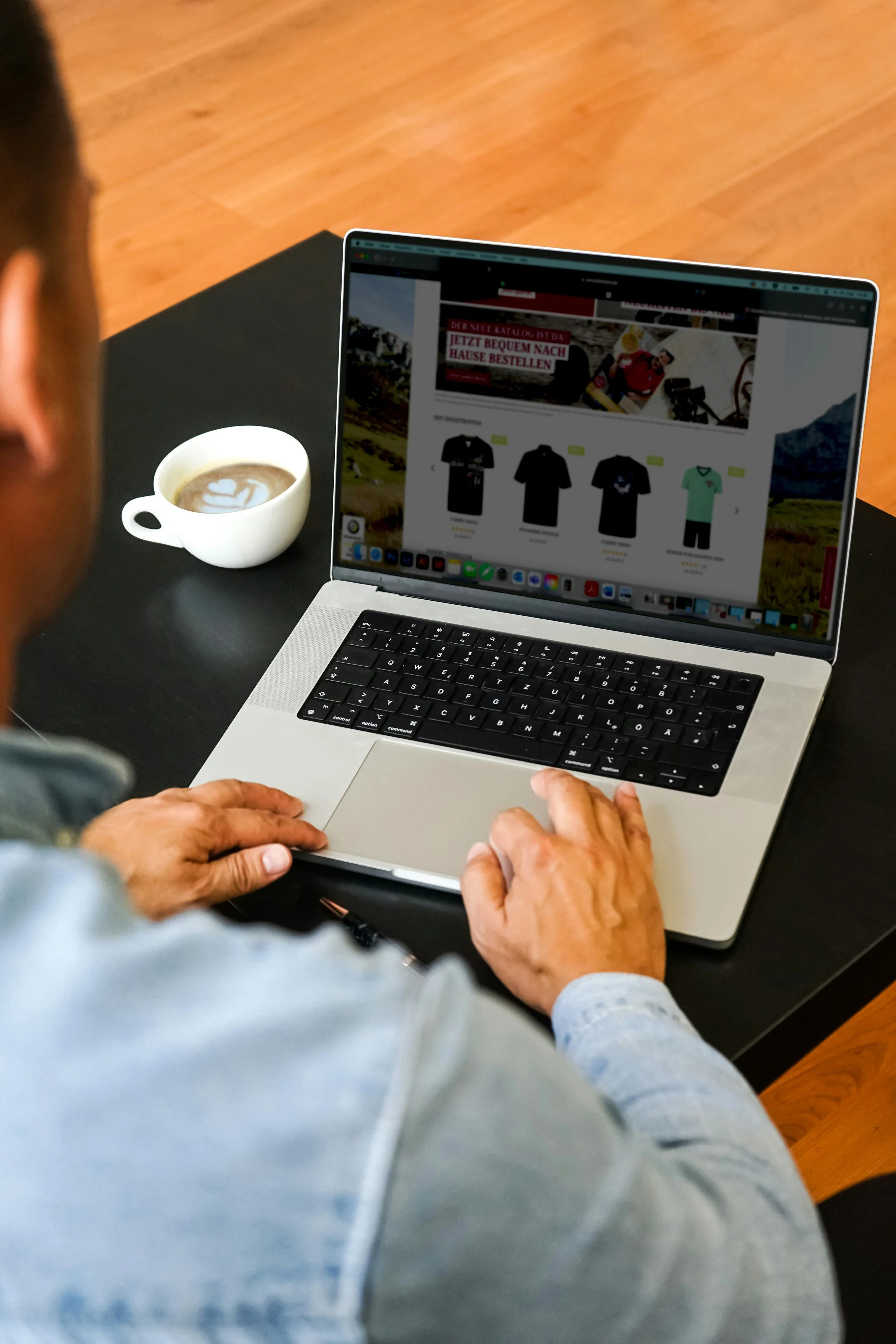 A person using a laptop on a black table, browsing an online store displaying various t-shirts and a green shirt. There is a cup of coffee with heart latte art on the table. The person has short hair and is wearing a denim shirt.