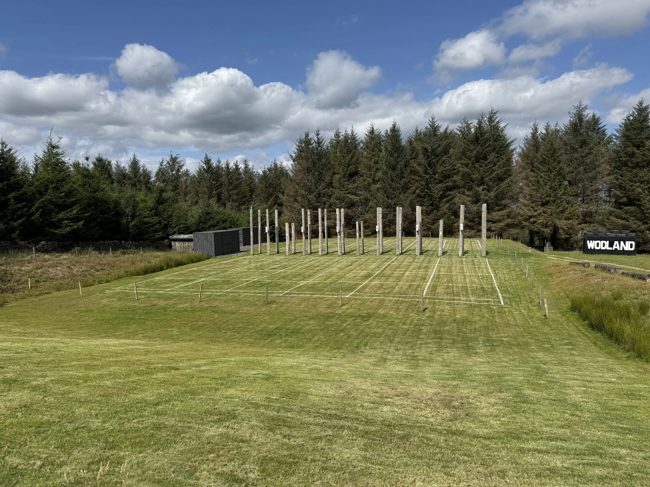 A grassy sports field with equipment poles and a sign that reads "WOLAND," surrounded by a forest of tall evergreen trees under a partly cloudy sky.