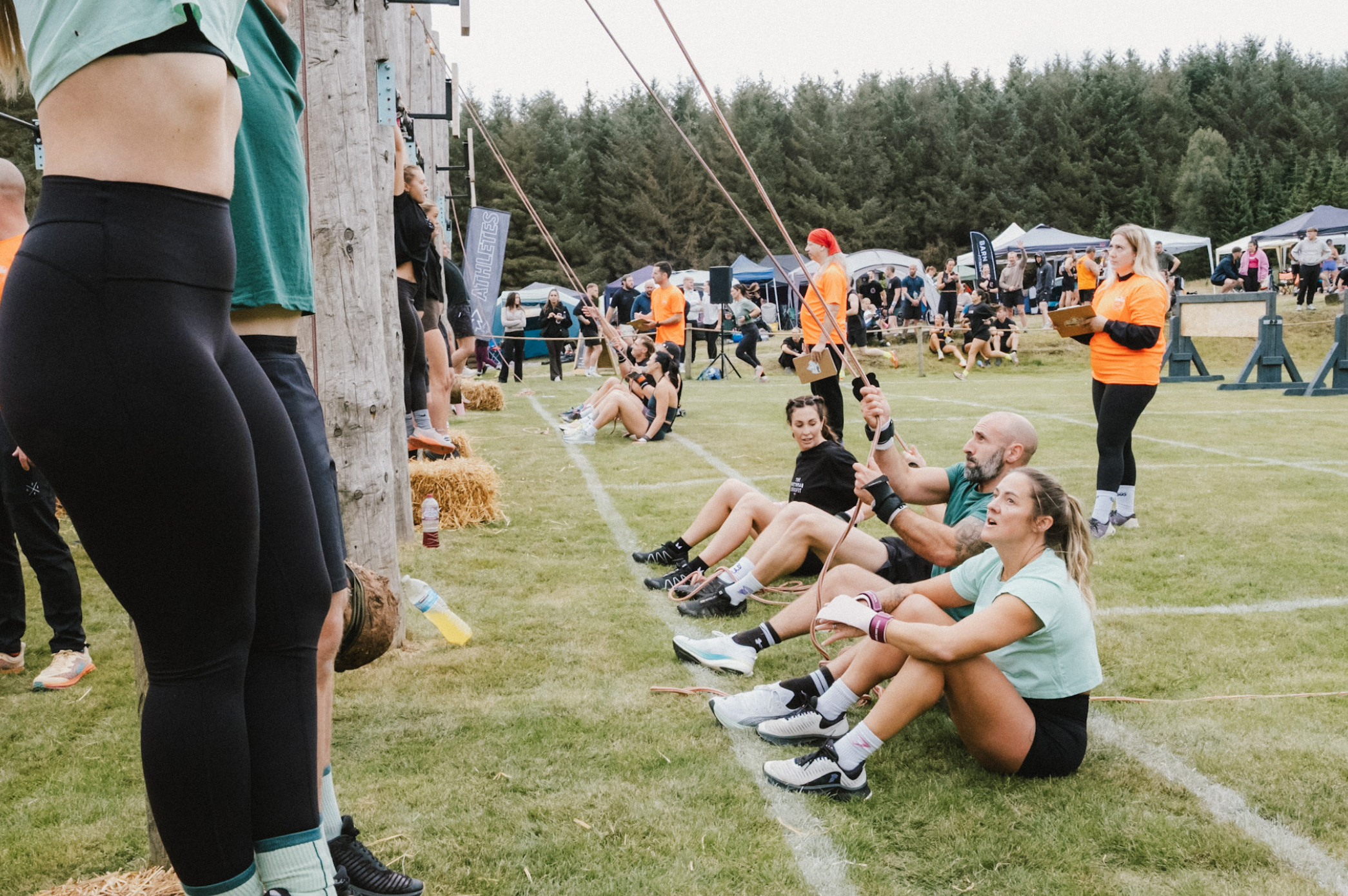 People sitting on the grass in front of an obstacle wall at an outdoor event, with others walking and tents in the background.