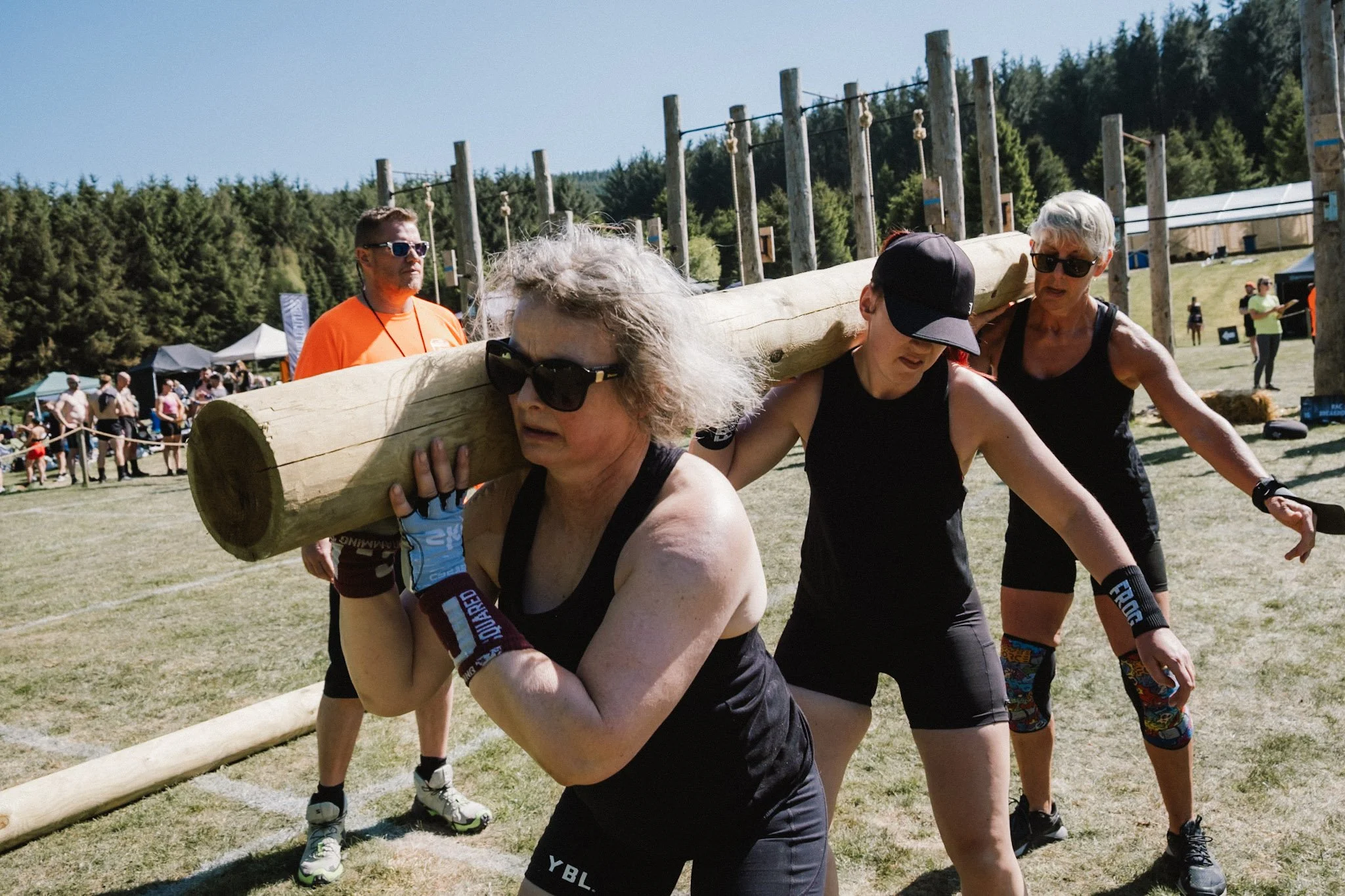 Women and men participating in a strongman competition outdoors, carrying a large log on their shoulders, with a crowd and trees in the background.