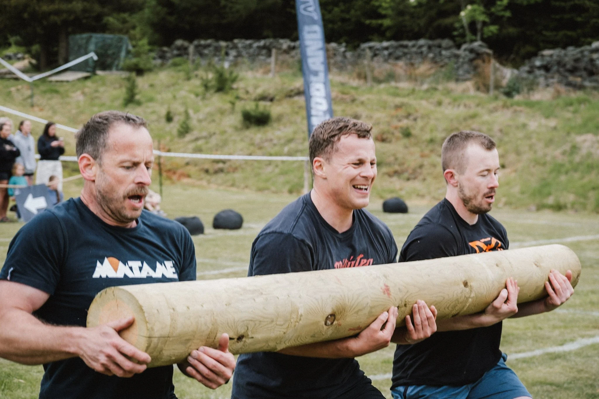 Three men participating in an outdoor strength competition, carrying a large log together during daytime in a grassy area with spectators in the background.