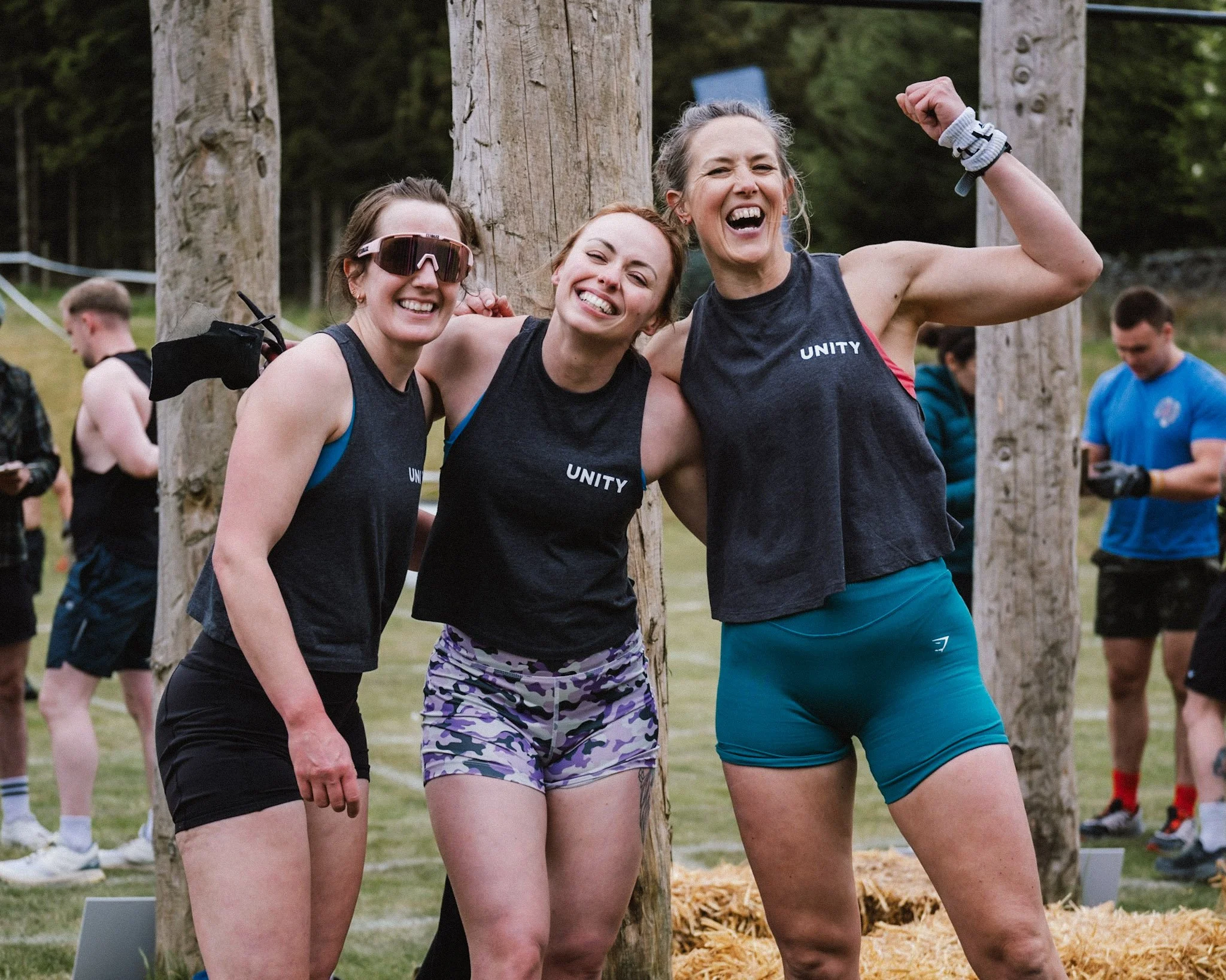 Three women standing together outdoors, smiling and showing off their muscles with one woman flexing her arm, during a recreational or sporting event with other people in the background.