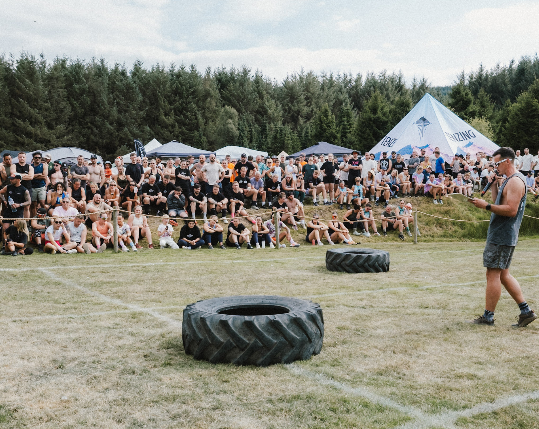 A large outdoor crowd of people gathered for an event, sitting on a grassy field behind a rope barrier, with several tents and a large mountain-shaped tent in the background. In the foreground, a person wearing sunglasses, a sleeveless shirt, and shorts is holding a microphone and looking at a phone. Two large tires are on the ground near the person.