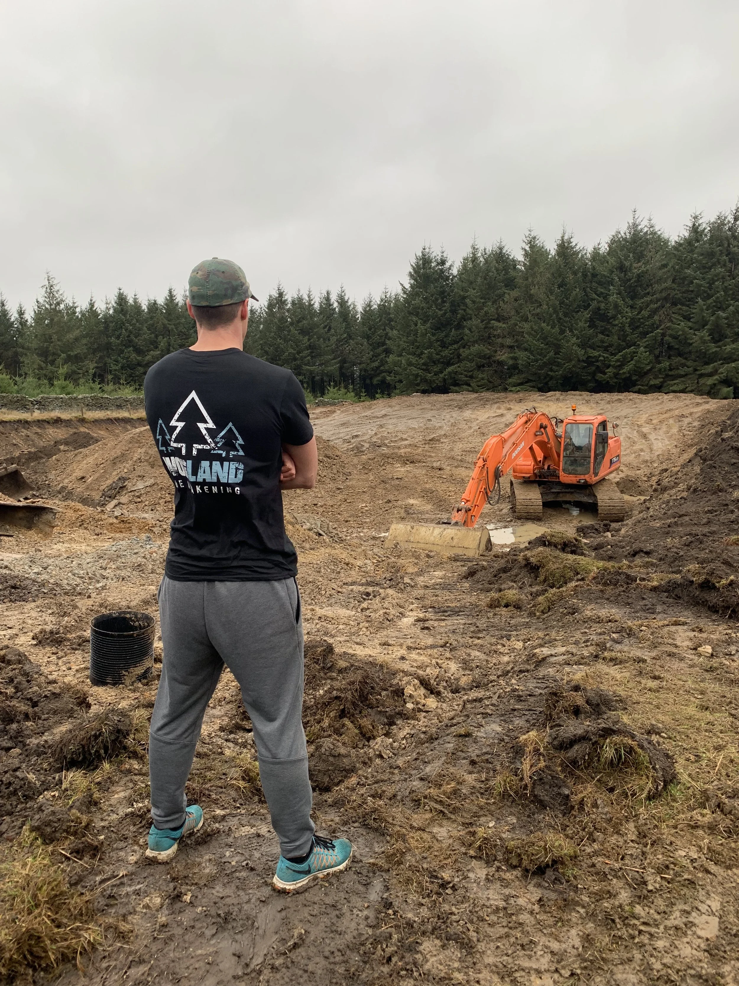 A person wearing a camouflage cap, black t-shirt, gray sweatpants, and teal running shoes stands on muddy ground with their back facing a small orange excavator, with a background of dense green trees under an overcast sky.