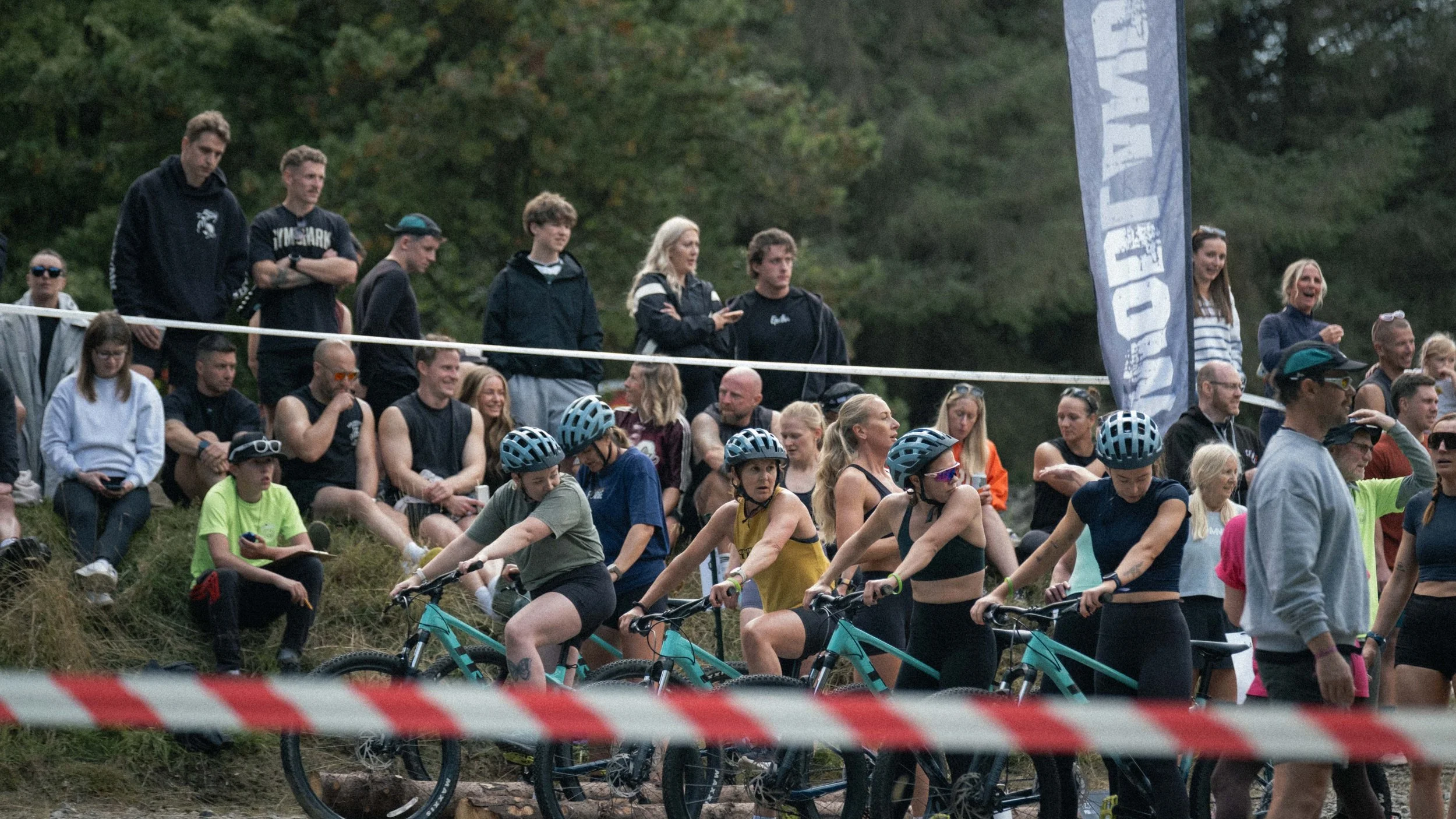 Group of people gathered at an outdoor event near a forest, some on bikes and some standing, with a flag displaying the text 'WAVE' in the background.