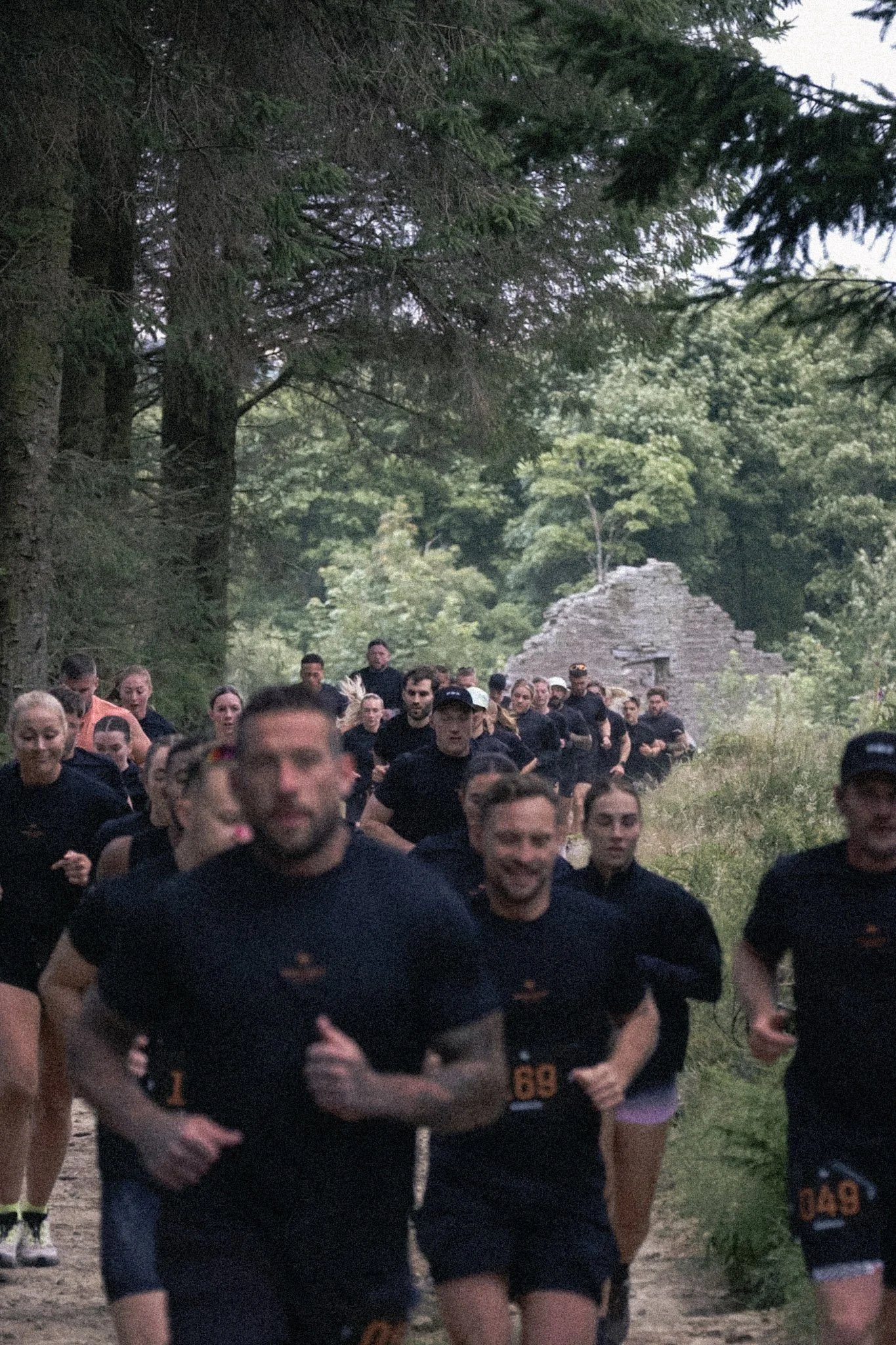 A large group of people running outdoors on a forest trail, surrounded by tall trees and greenery, with an old stone structure in the background.