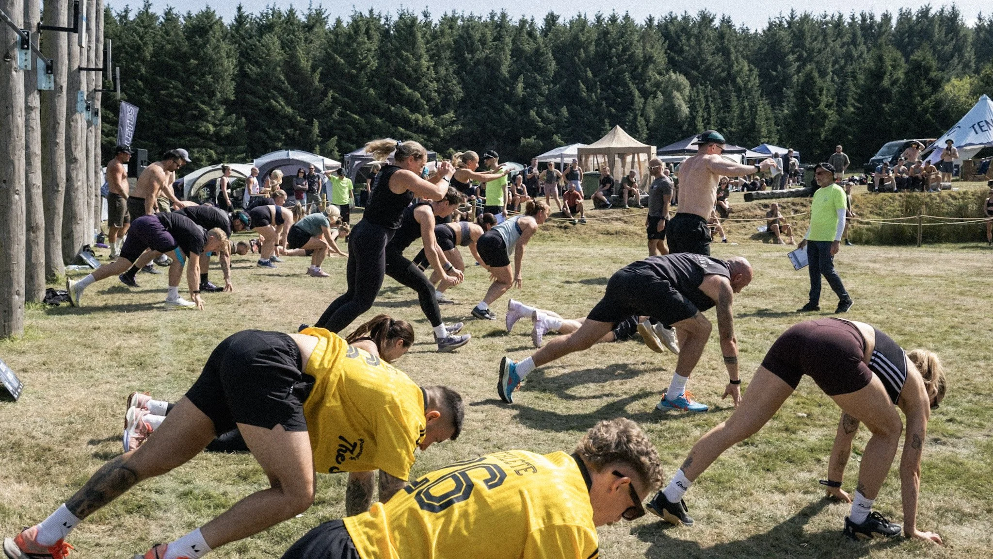 Group of people stretching and warming up outdoors on a grassy field, with tents and trees in the background, during a sunny day.