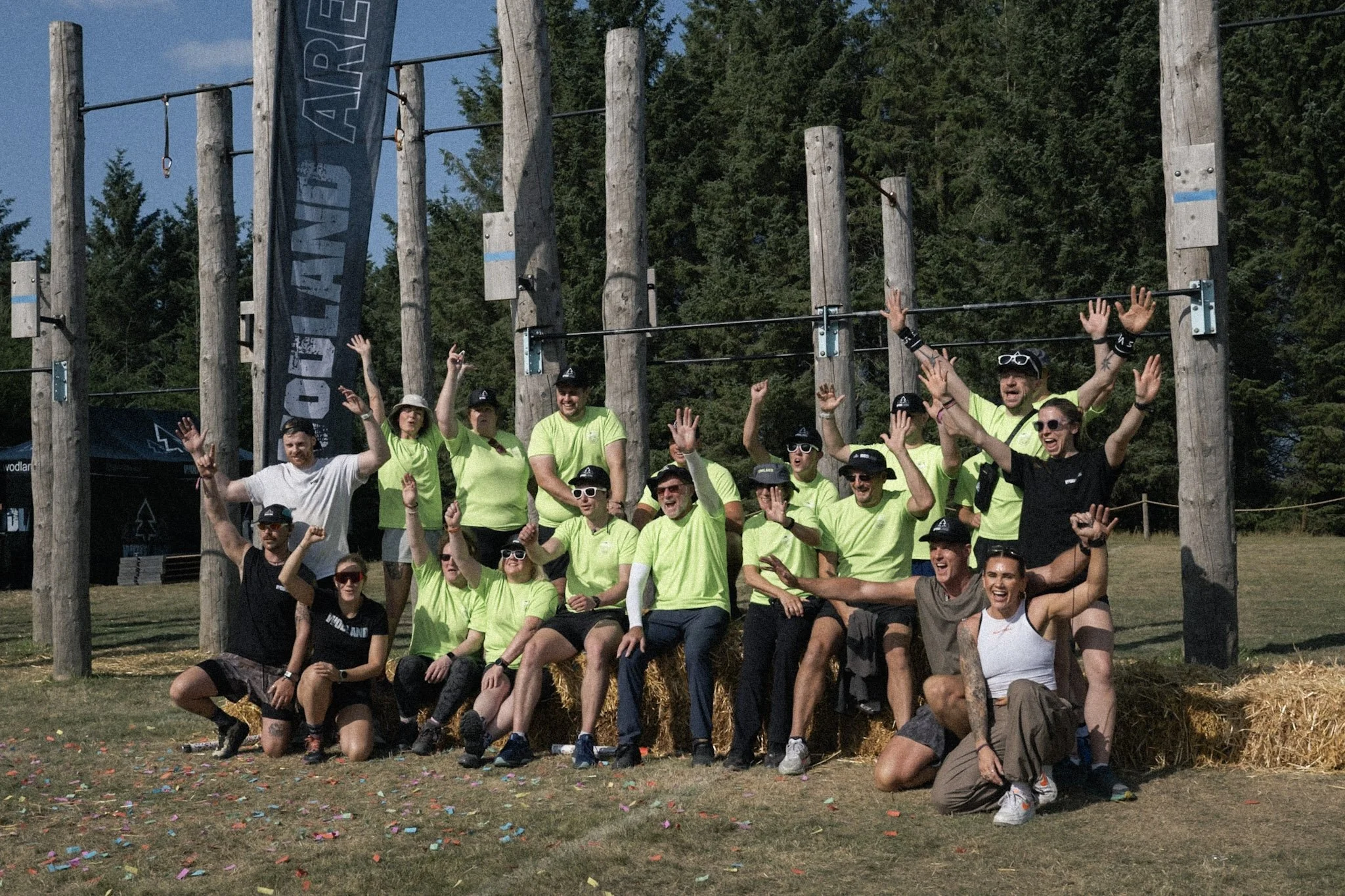 Group of people posing outdoors in front of wooden climbing structures on a sunny day, some wearing bright yellow shirts, with confetti on the ground.