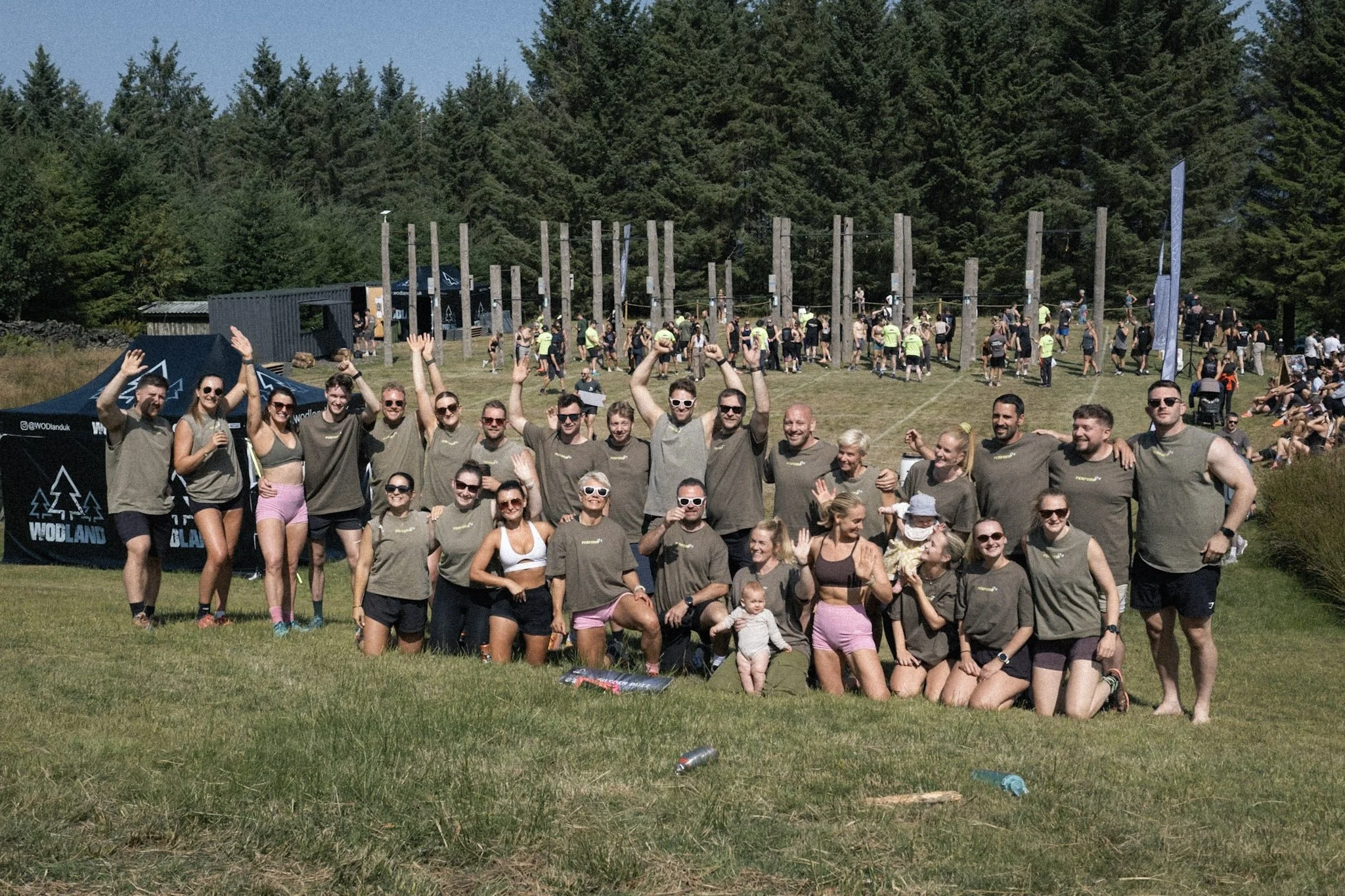 A large group of people gathered outdoors on a sunny day, some wearing matching shirts, posing for a photo with a forested background and an obstacle course, with children and spectators nearby.