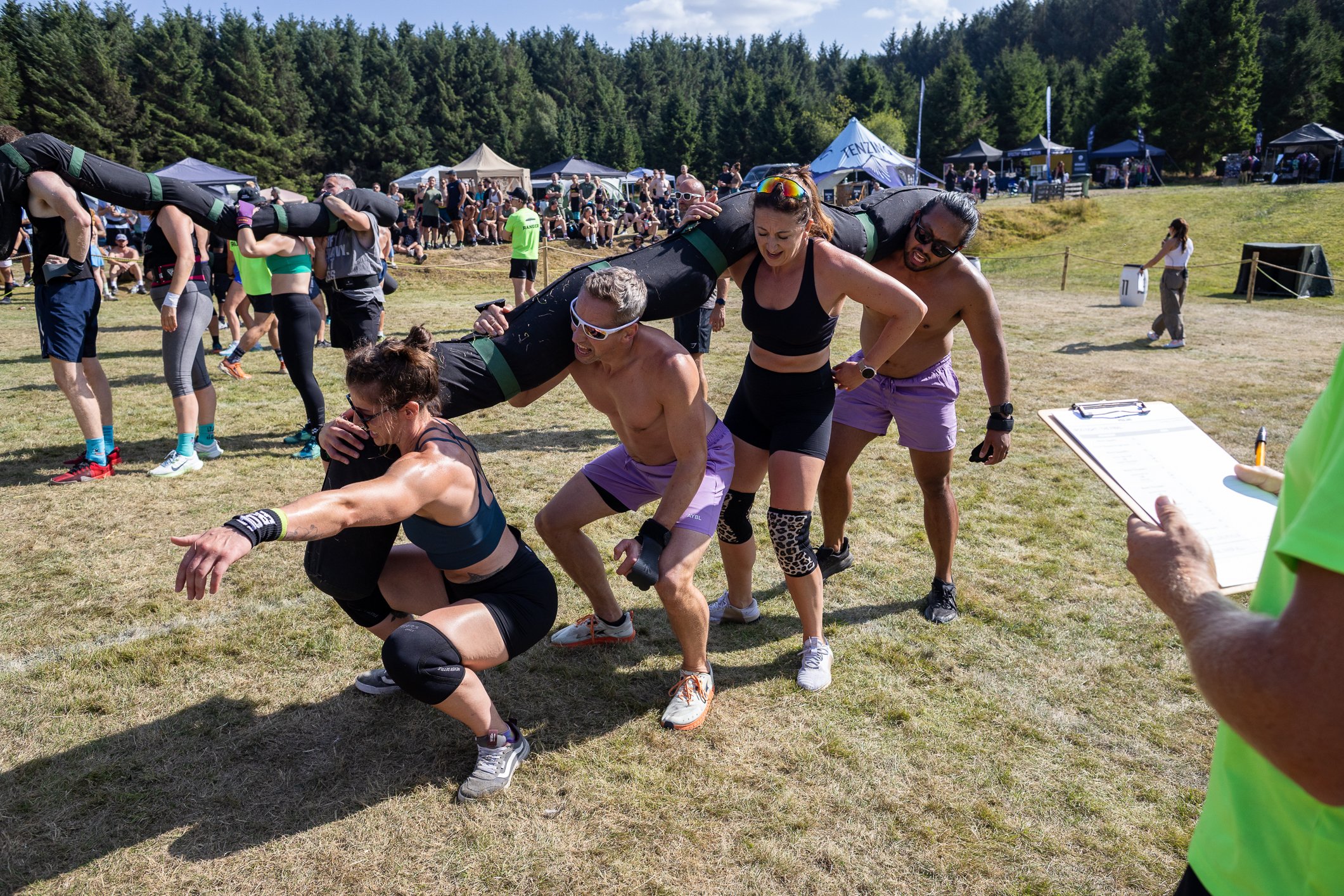 People participating in an outdoor team-building activity, forming a human pyramid on a grassy field with tents and spectators in the background.