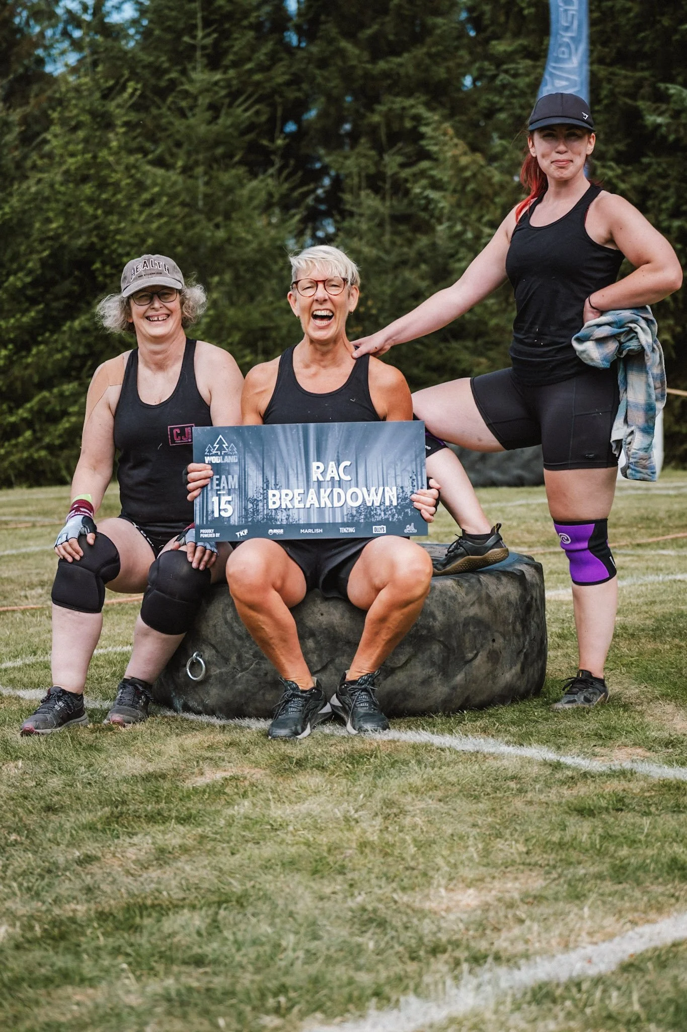 Three women posing outdoors on a grassy field during daytime, with a wooded background, holding a sign that reads 'RAC Breakdown' and 'Team 15', wearing black athletic outfits and knee braces, one woman wearing a hat, and one woman standing with her leg on a large rock.