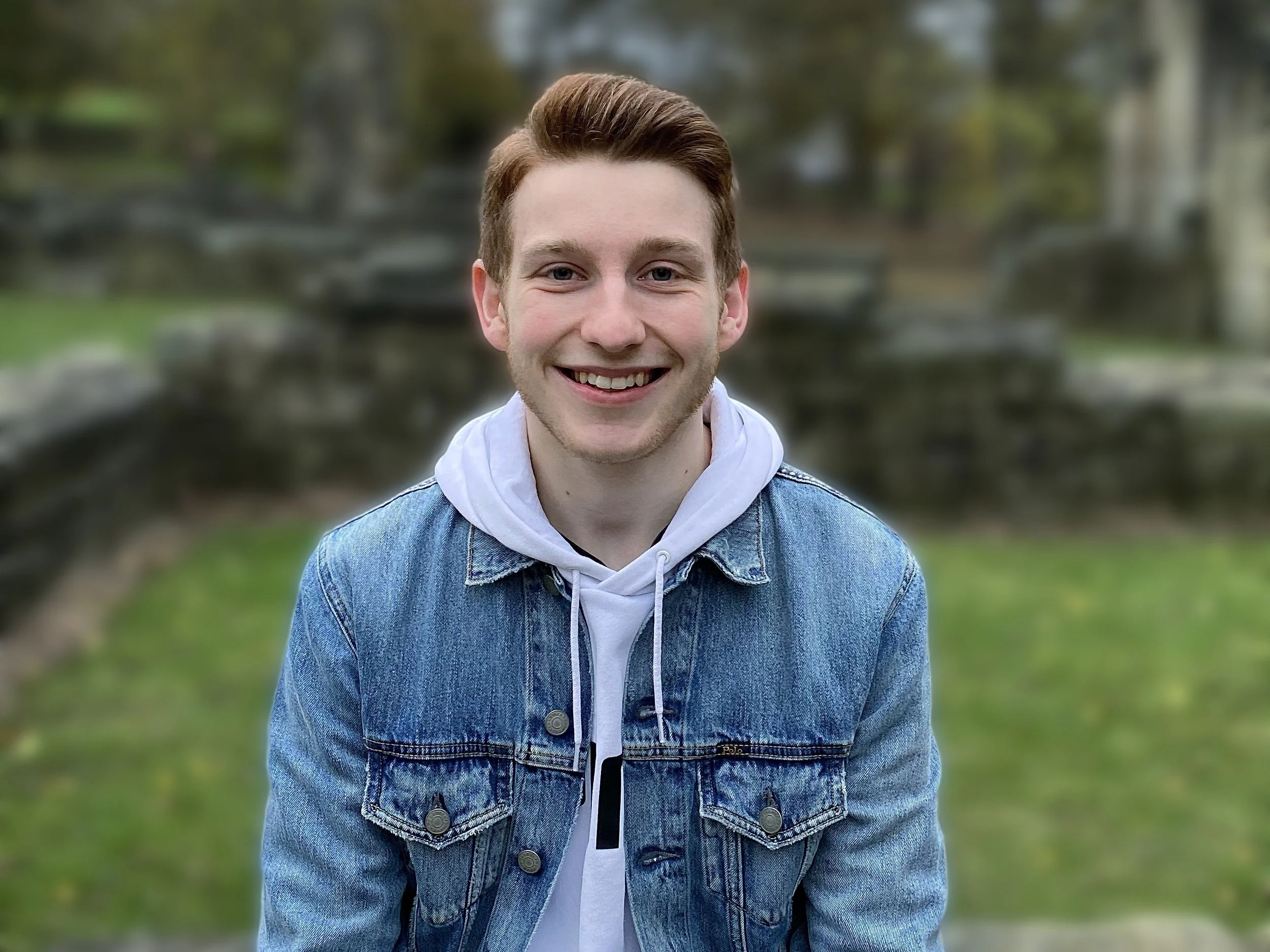 A young man with styled brown hair, smiling, wearing a white hoodie layered under a blue denim jacket, outdoors with greenery and blurred trees in the background.