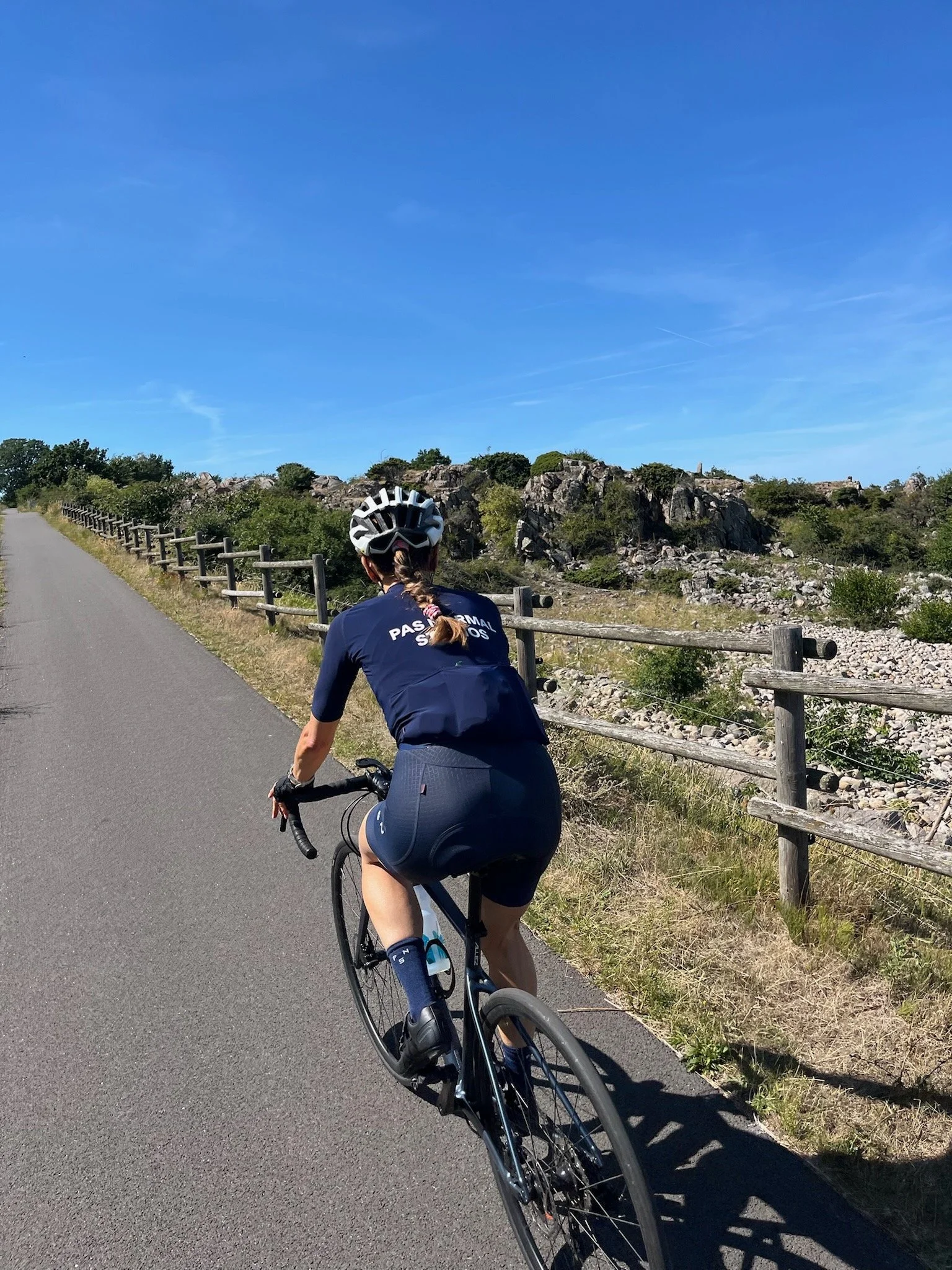 En kvinde cykler på en vej omgivet af klipper og grøn vegetation under en klar blå himmel.