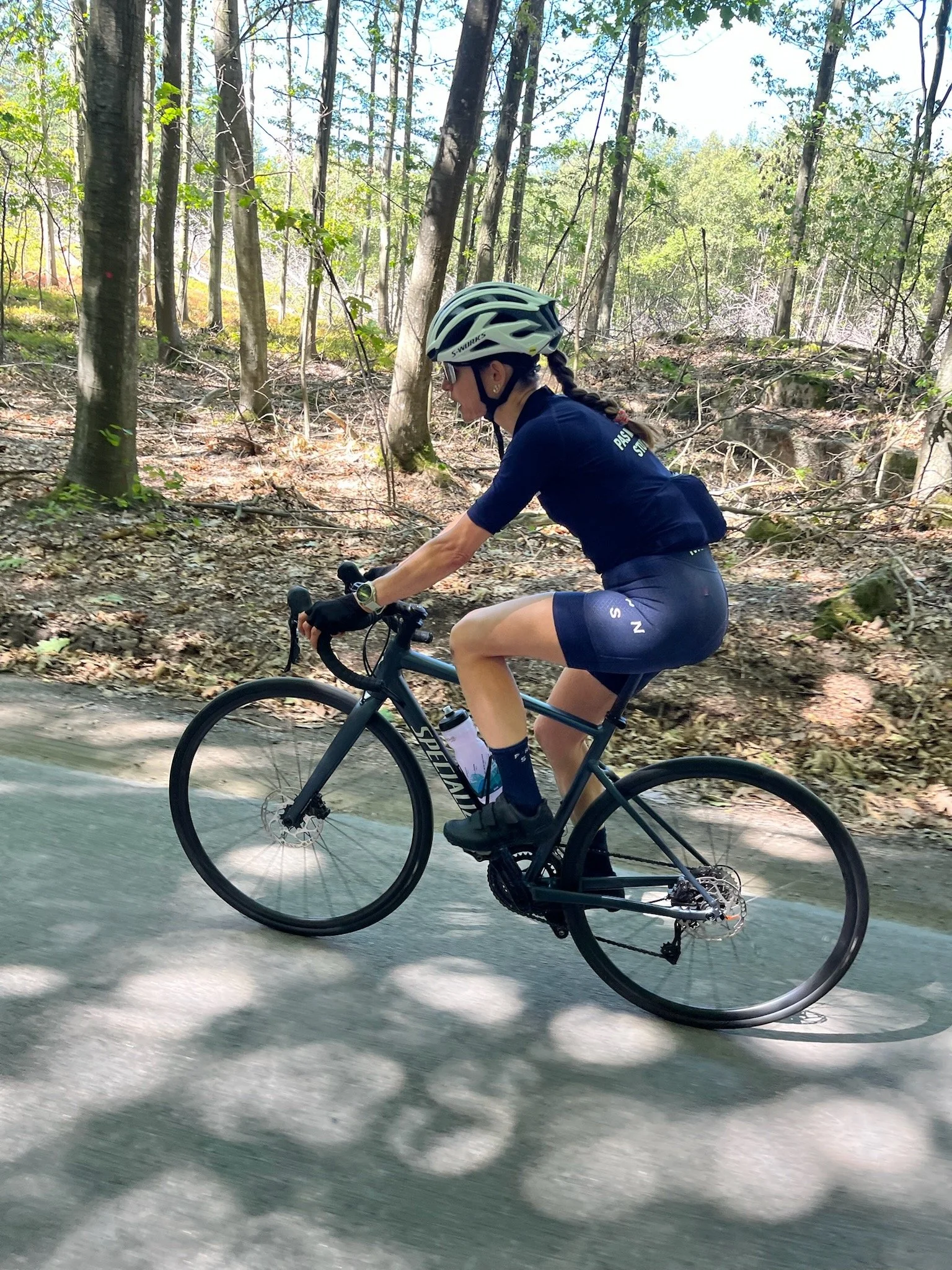 En kvinde cykler på en landvej gennem en skov med grønne træer og sollys, iført cykelhjelm og sportsudstyr.