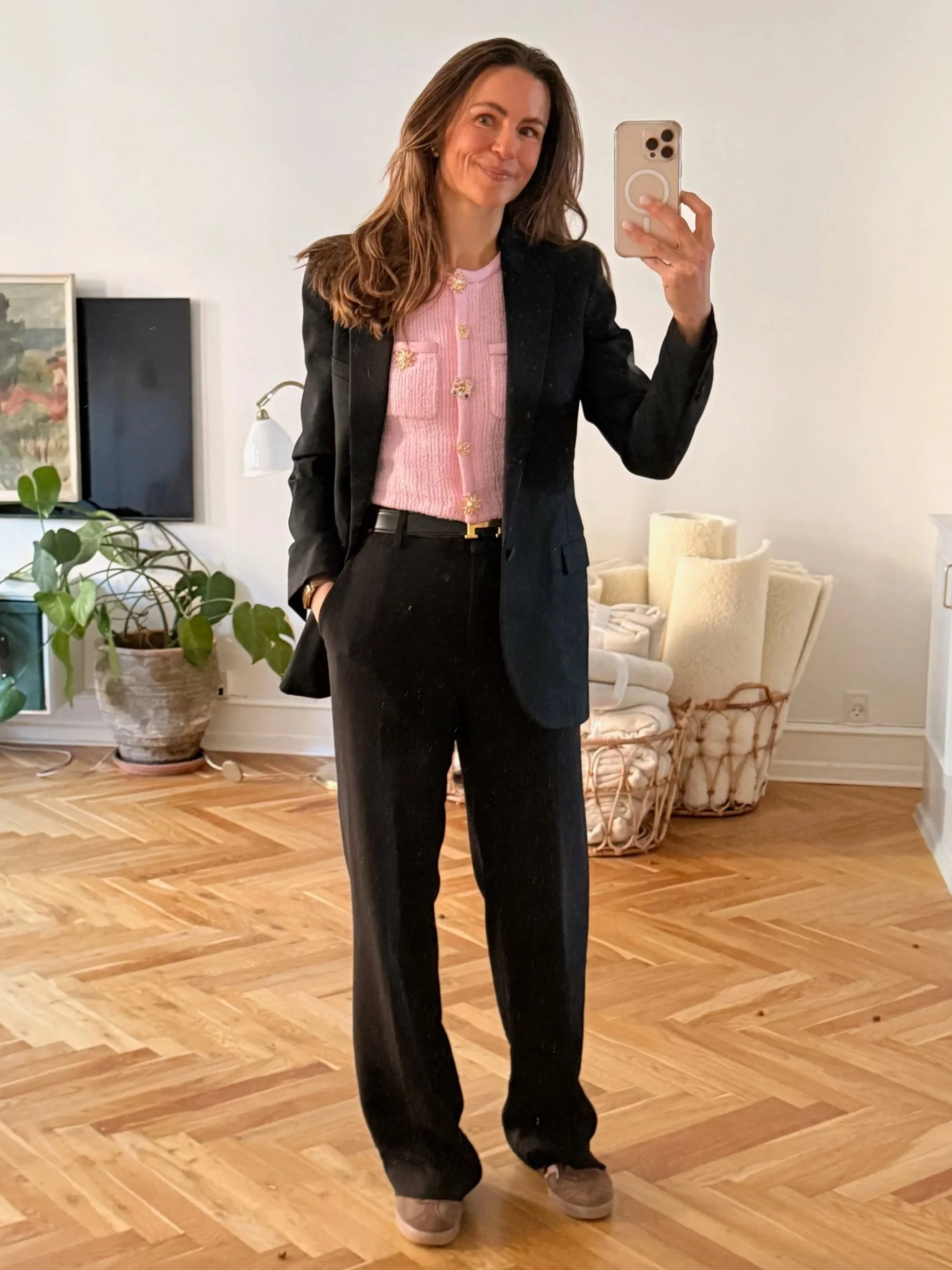 En person tager et selfie i en stue med trægulv, der er pyntet med planter, malerier og moderne møbler, og personen har på en sort blazer, pink sweater med guldknapper og sorte bukser.