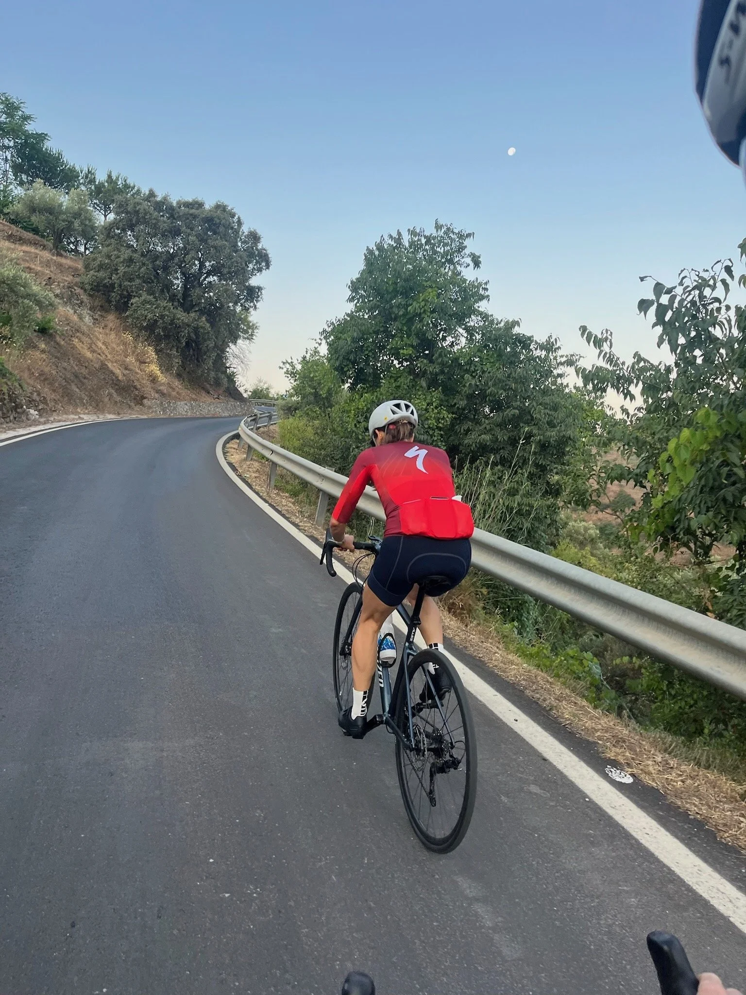 To cyklister på en snoet bjergvej om aftenen, den ene i en rød cykeltrøje og hjelm. Der er grønt løv og en blå himmel med månen i baggrunden.