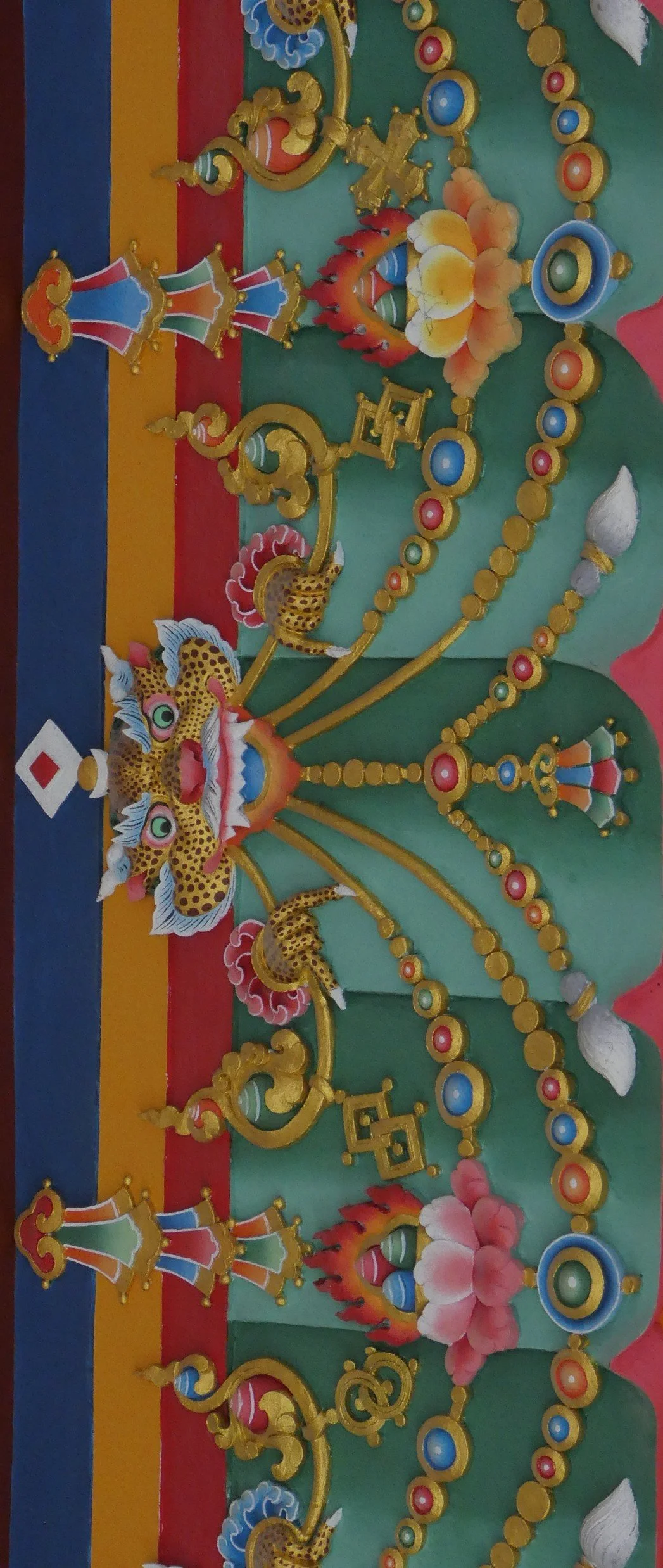 Colorful decorative sculpture featuring a central creature with a feline face and multiple golden fin-like extensions, surrounded by floral elements, beads, and structures with a rainbow-striped background.