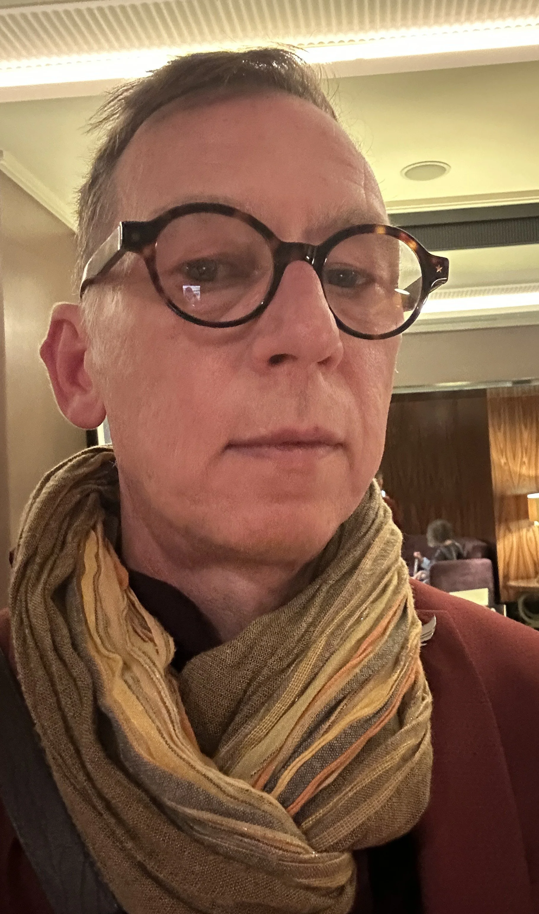 A person with short brown hair, wearing round black glasses, a striped scarf, and a maroon sweater, sitting in a room with wooden wall panels.