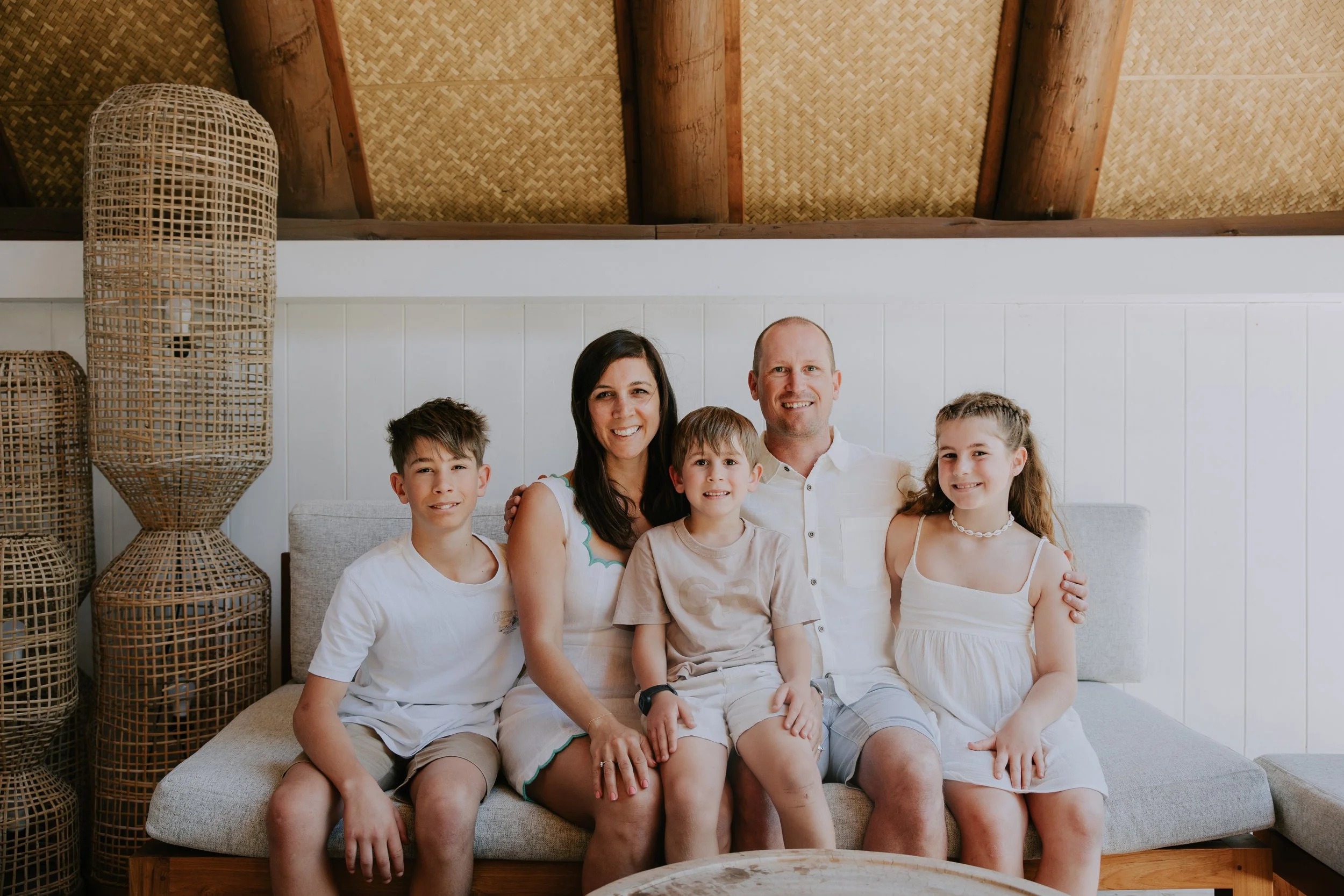 A family of five sitting together on a sofa in a room with woven and wooden decor, smiling at the camera.