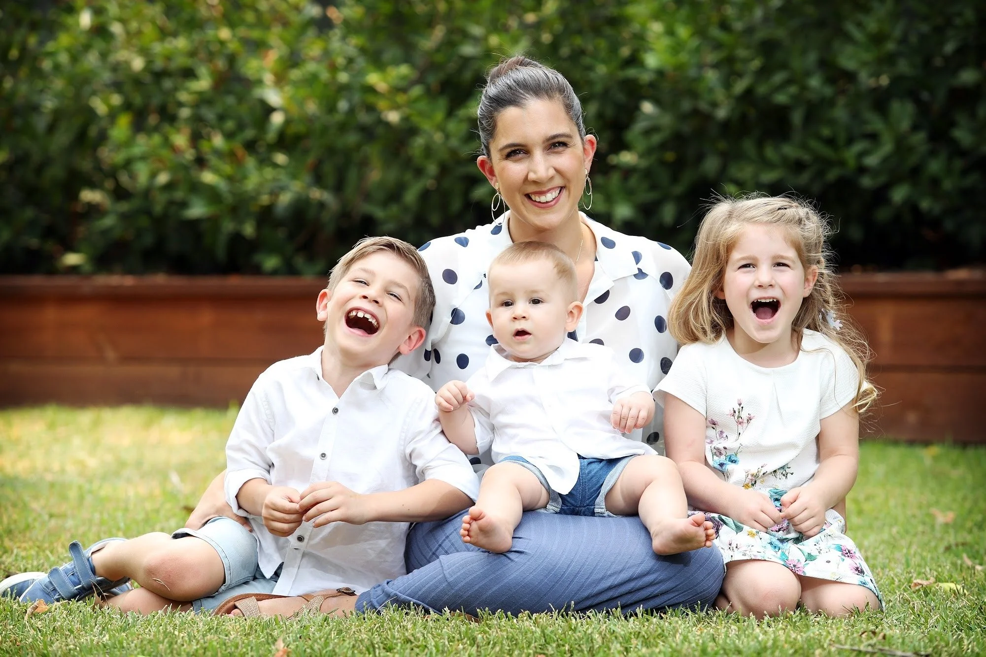A smiling woman with three young children sitting on grass in a park, with greenery and trees in the background.