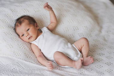 Baby lying on a white textured blanket, wearing a white sleeveless outfit.