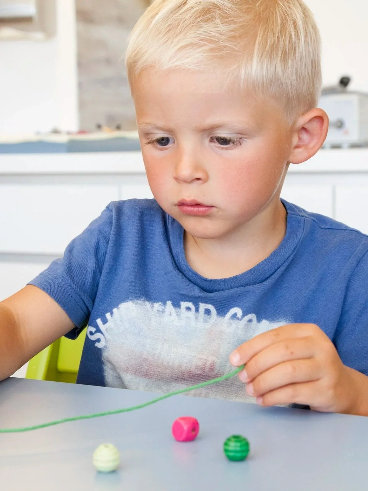 Un jeune garçon blond concentré joue avec de la laine et des boutons colorés sur une table.
