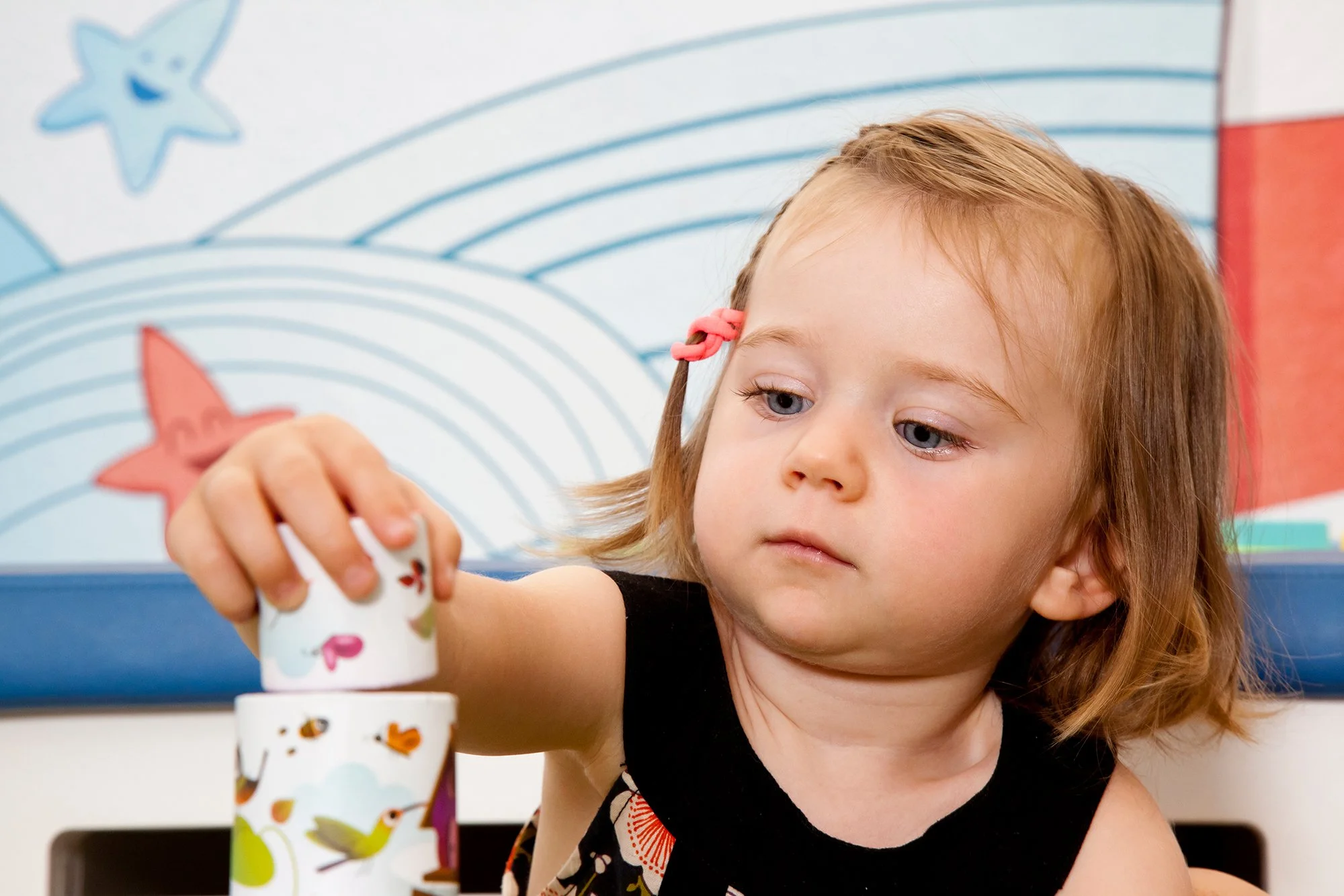 Une jeune fille aux cheveux roux et yeux bleus joue à un jeu de société avec des gobelets décorés d'animaux, dans une pièce avec un fond mural à rayures et étoiles de couleur bleue et rouge.