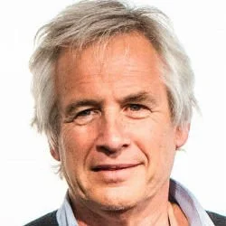 A middle-aged man with short gray hair and beard, wearing a gray blazer over a black plaid shirt, smiling against a plain white wall.